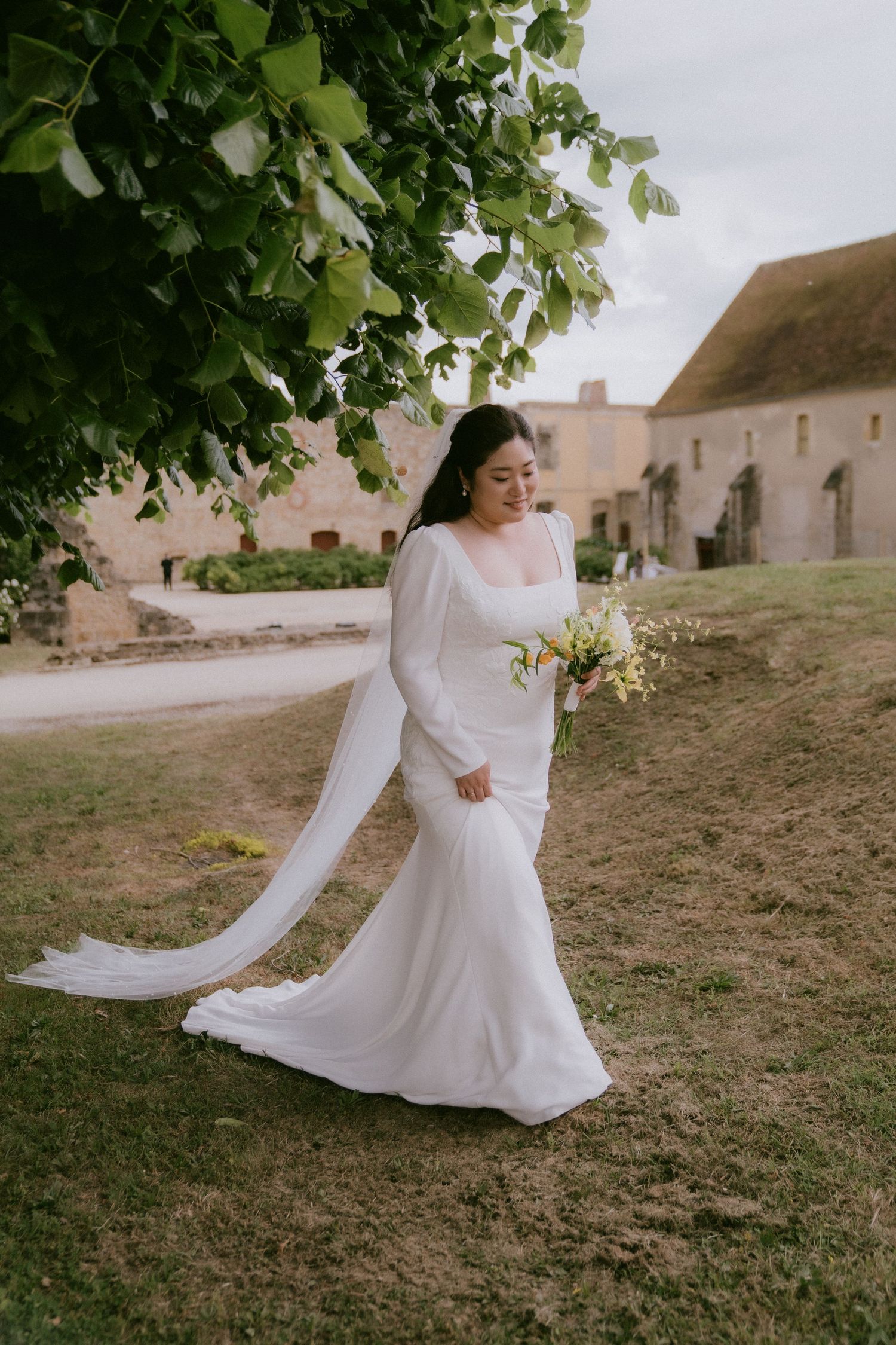 A person in a flowing white wedding dress twirls gracefully on a grassy hillside near historic buildings.