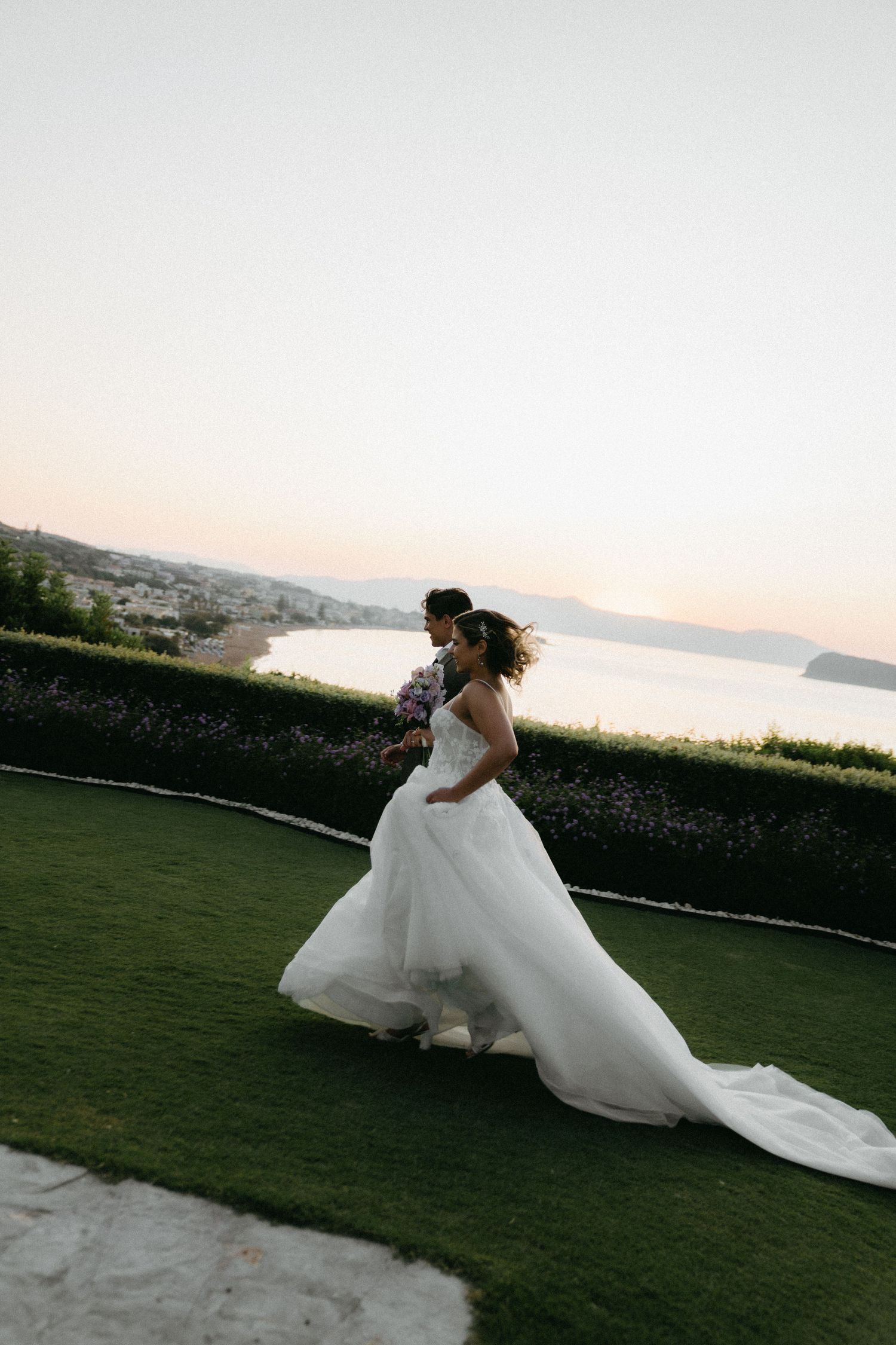 A couple in wedding attire dances together on a manicured lawn overlooking a scenic coastal view at sunset.