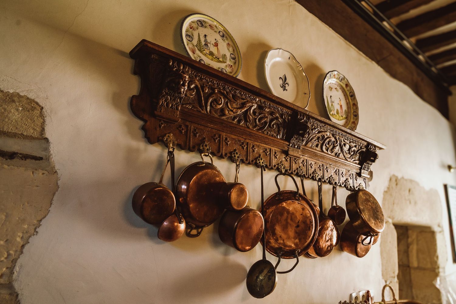 Rustic copper pots and pans hang from an ornate wooden rack beneath decorative plates on a white kitchen wall.
