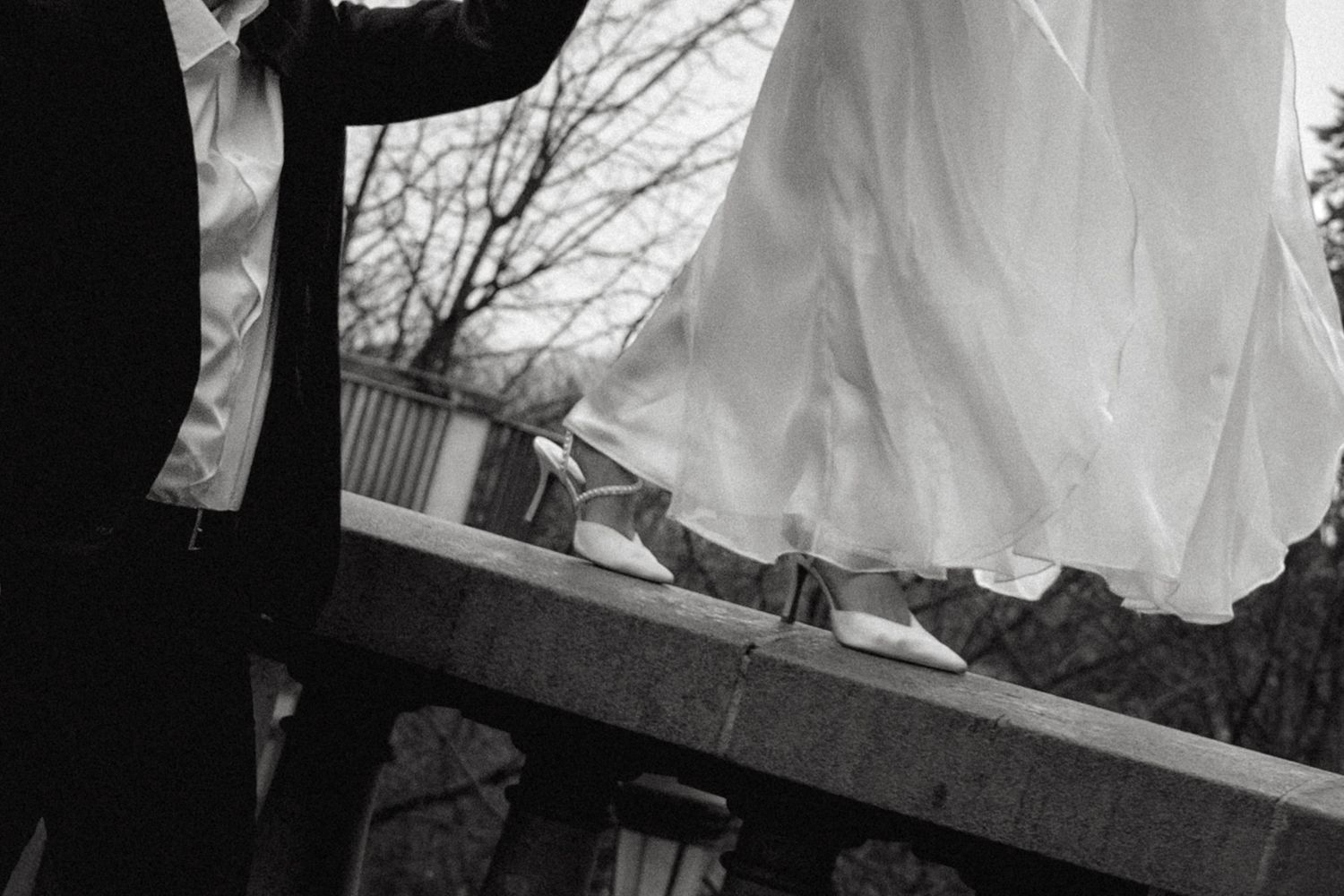 Black and white photo showing the hem of a flowing wedding dress and dress shoes on a wooden deck.