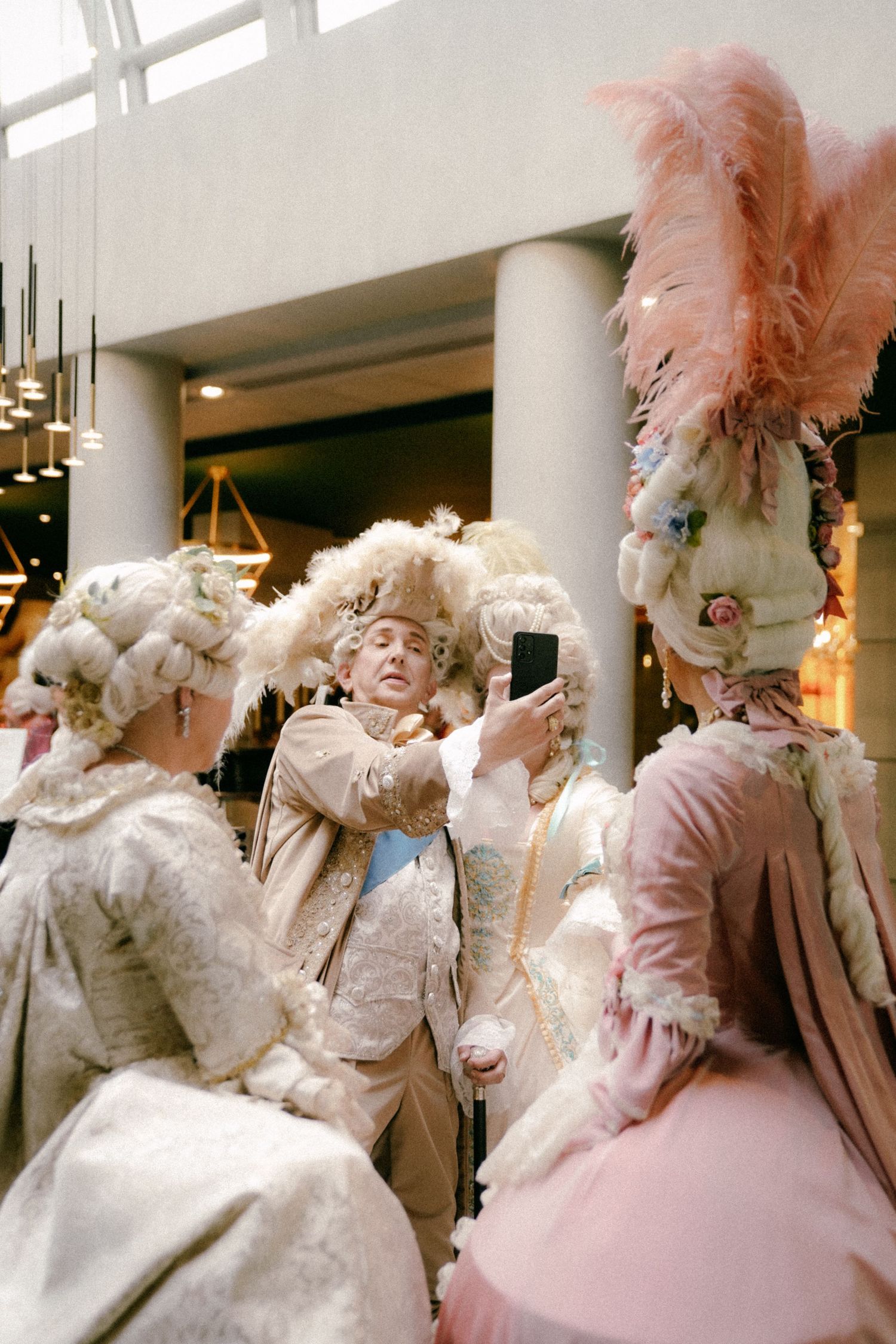 People in elaborate 18th century Rococo costumes with feathered masks and pastel pink and white outfits pose together.