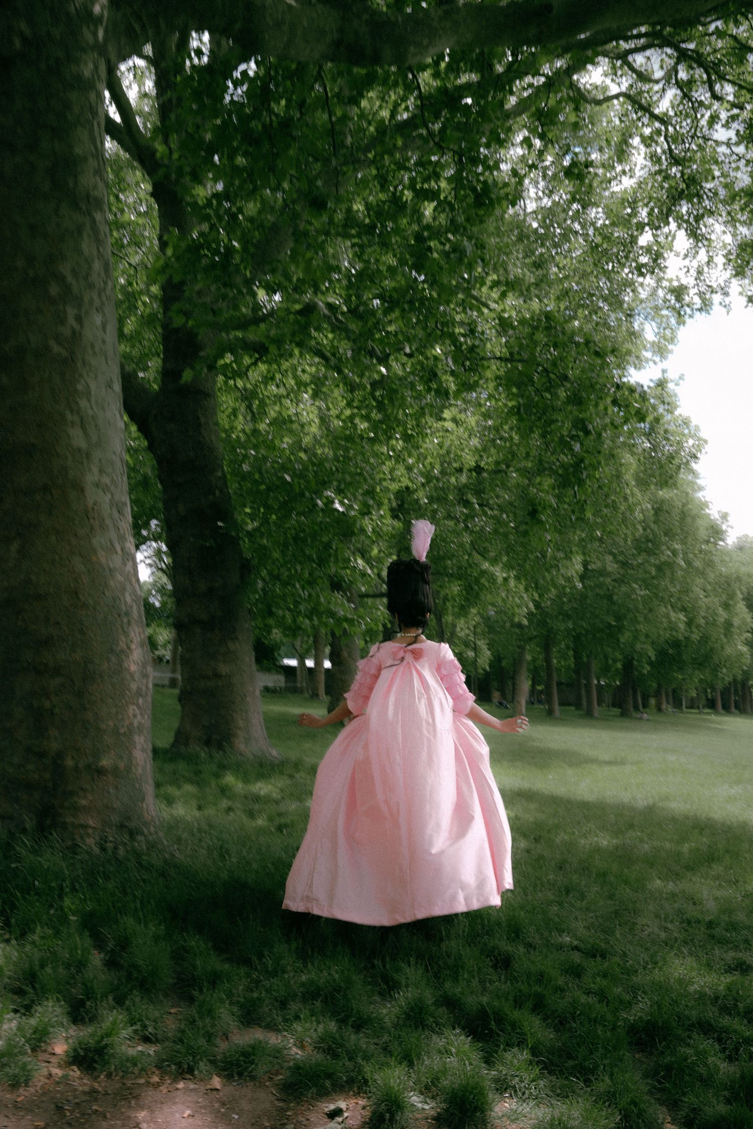 A pink Victorian-style dress displayed among tall trees in a serene park setting.