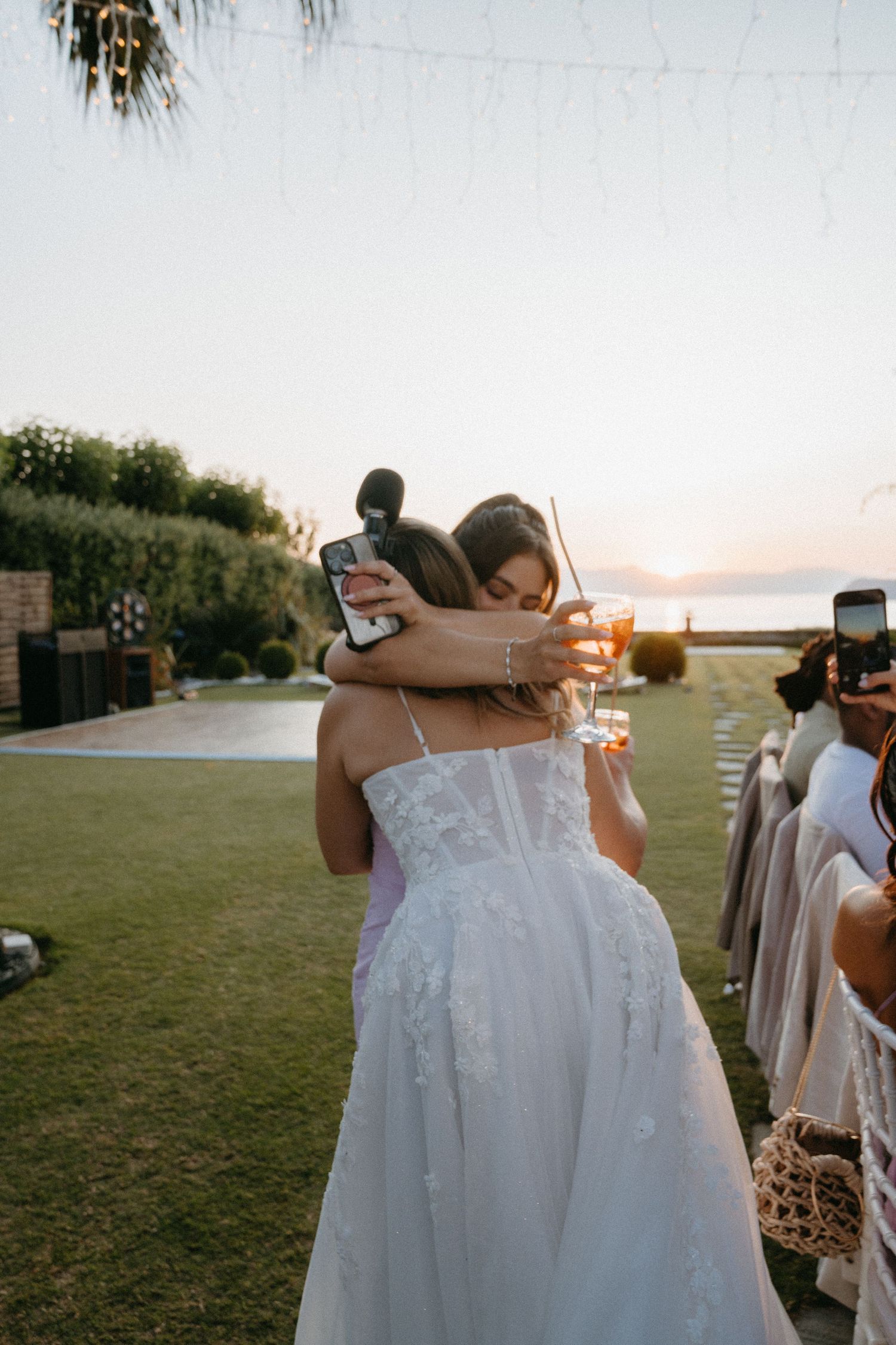 A person in a white wedding dress stands against a sunset backdrop while others take photos.