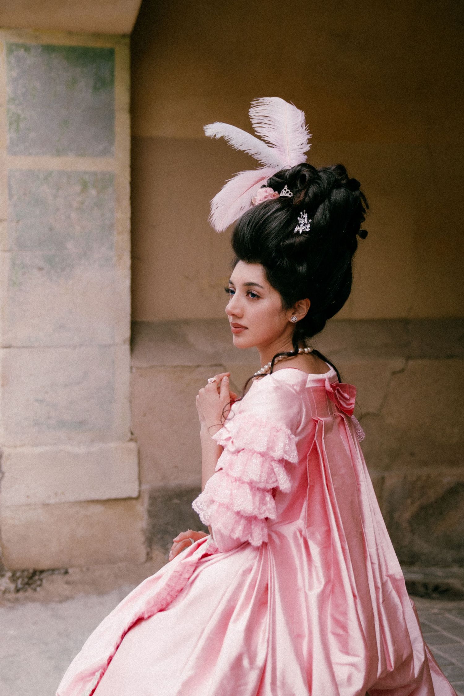 A person in a flowing pink Victorian-style dress with ruffles poses against a stone wall backdrop.