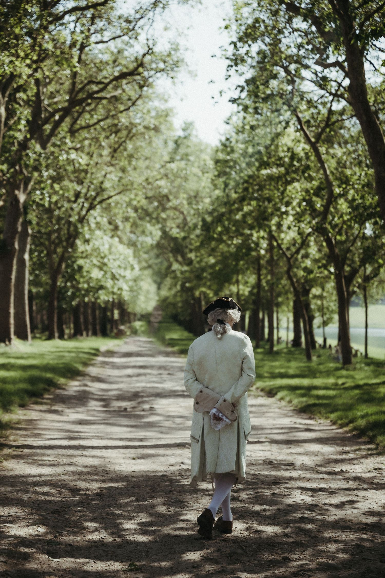 Person in 18th-century costume walking along a tree-lined path in a lush green park.