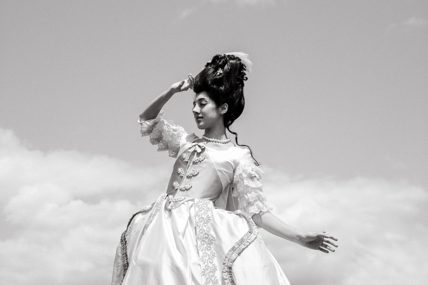 A person in an elegant white Victorian-style dress poses dramatically against a cloudy sky in black and white.