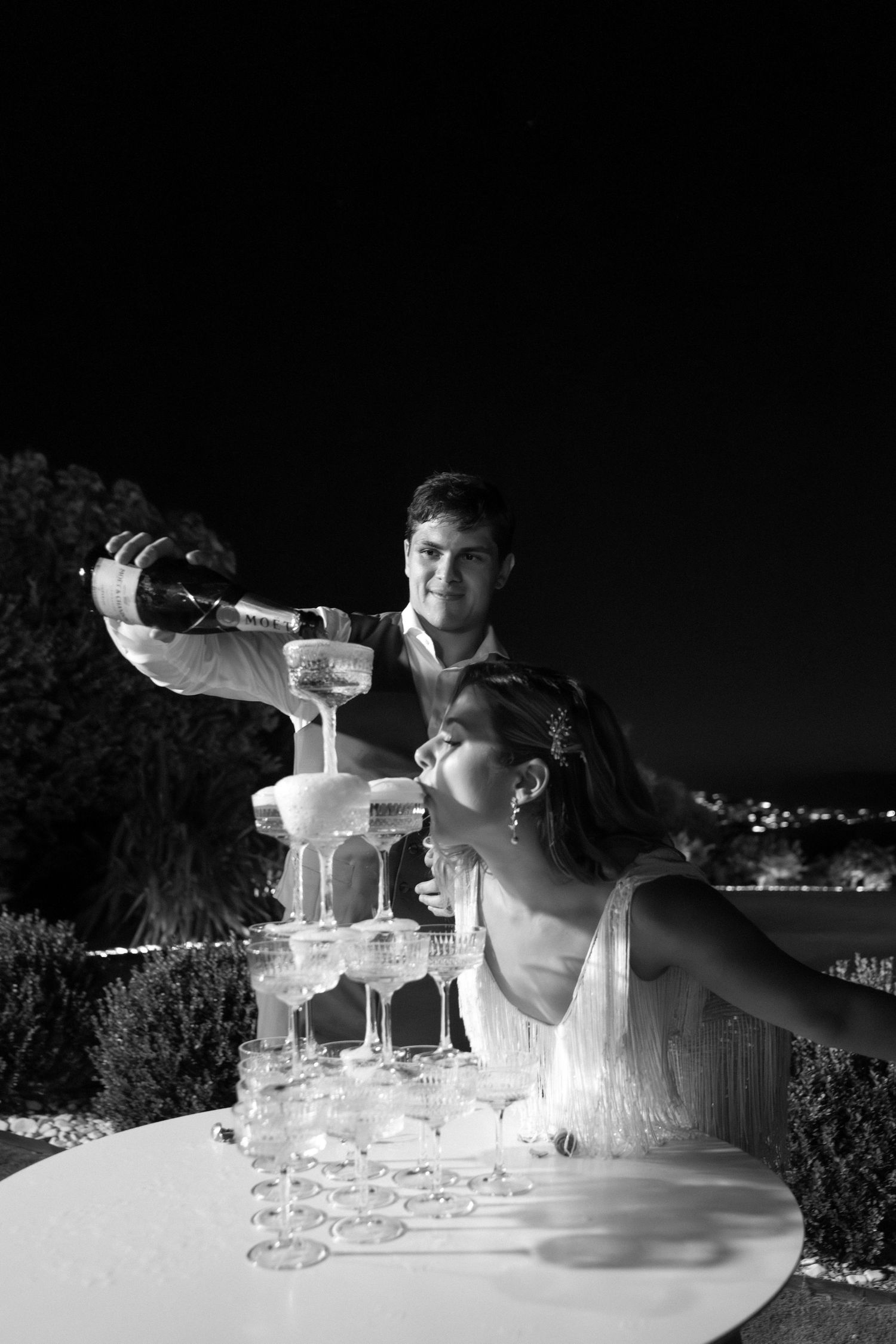 A champagne tower being filled at an elegant nighttime celebration with city lights in the background.