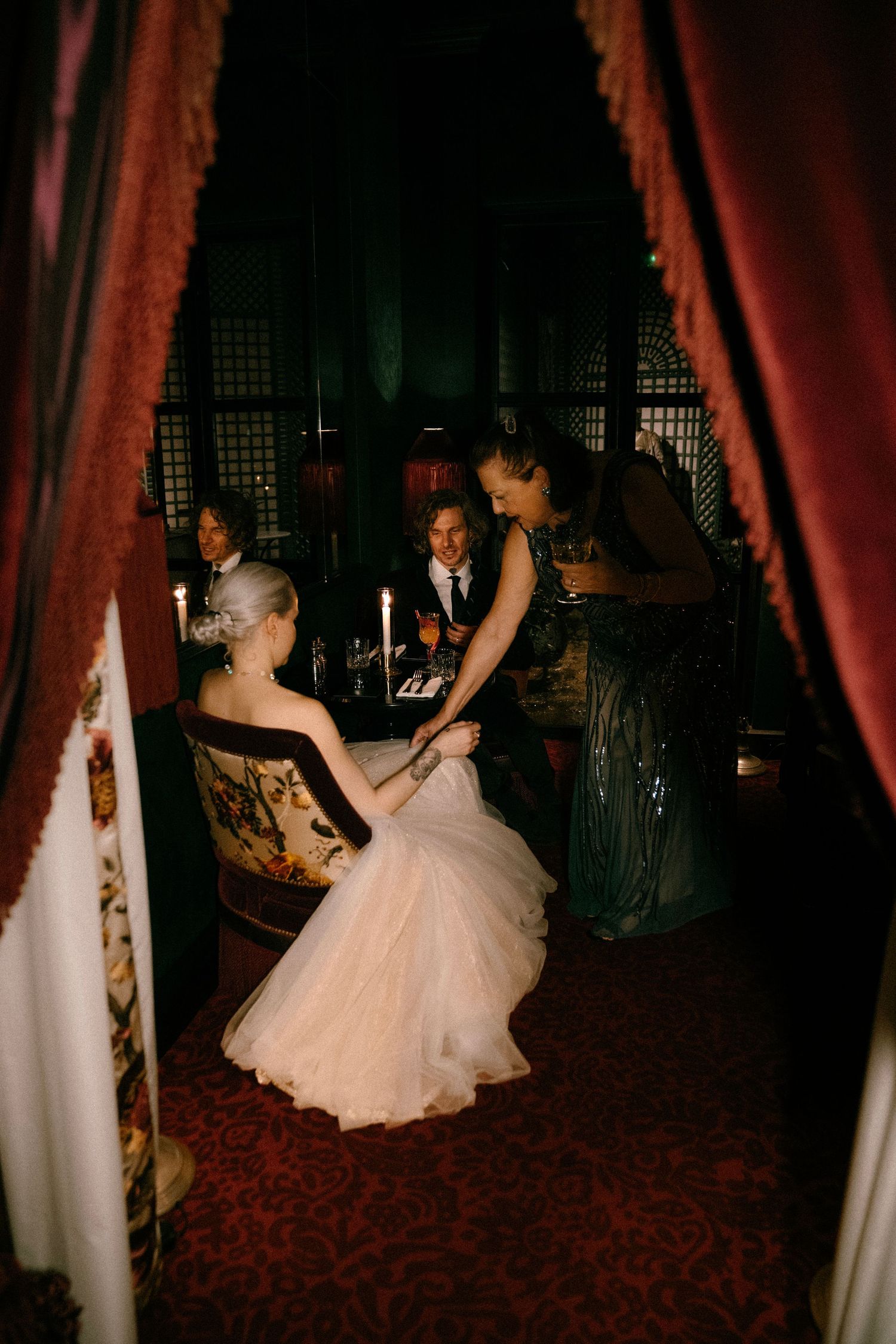 Candlelit scene through red curtains showing formal evening gathering in vintage setting.