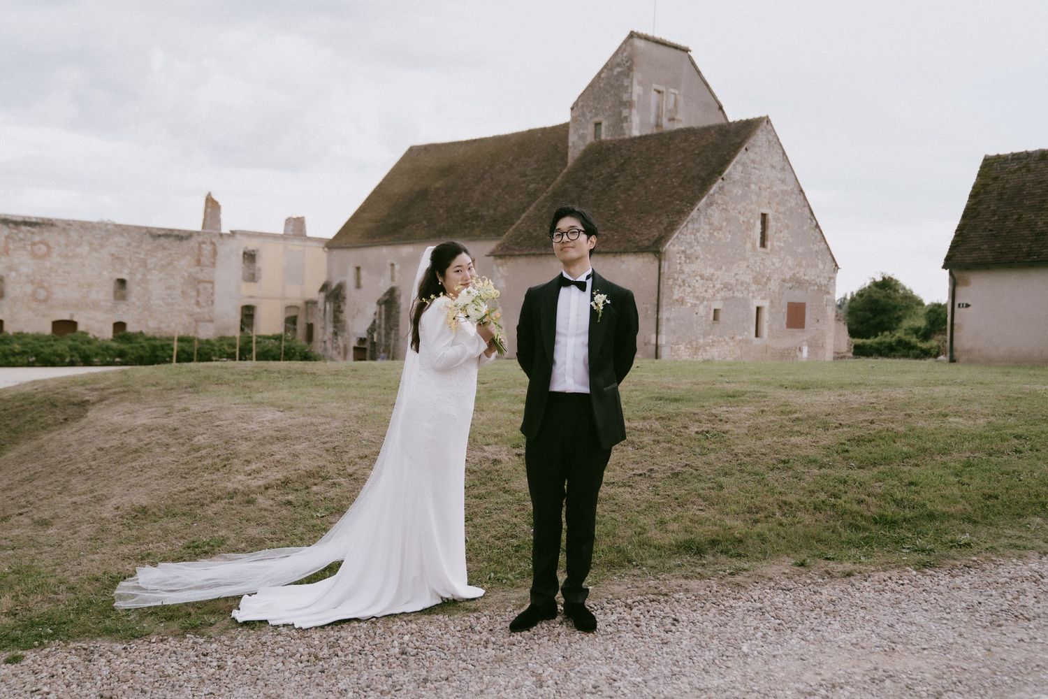 A romantic wedding couple stands in front of stone buildings at a rustic countryside venue.