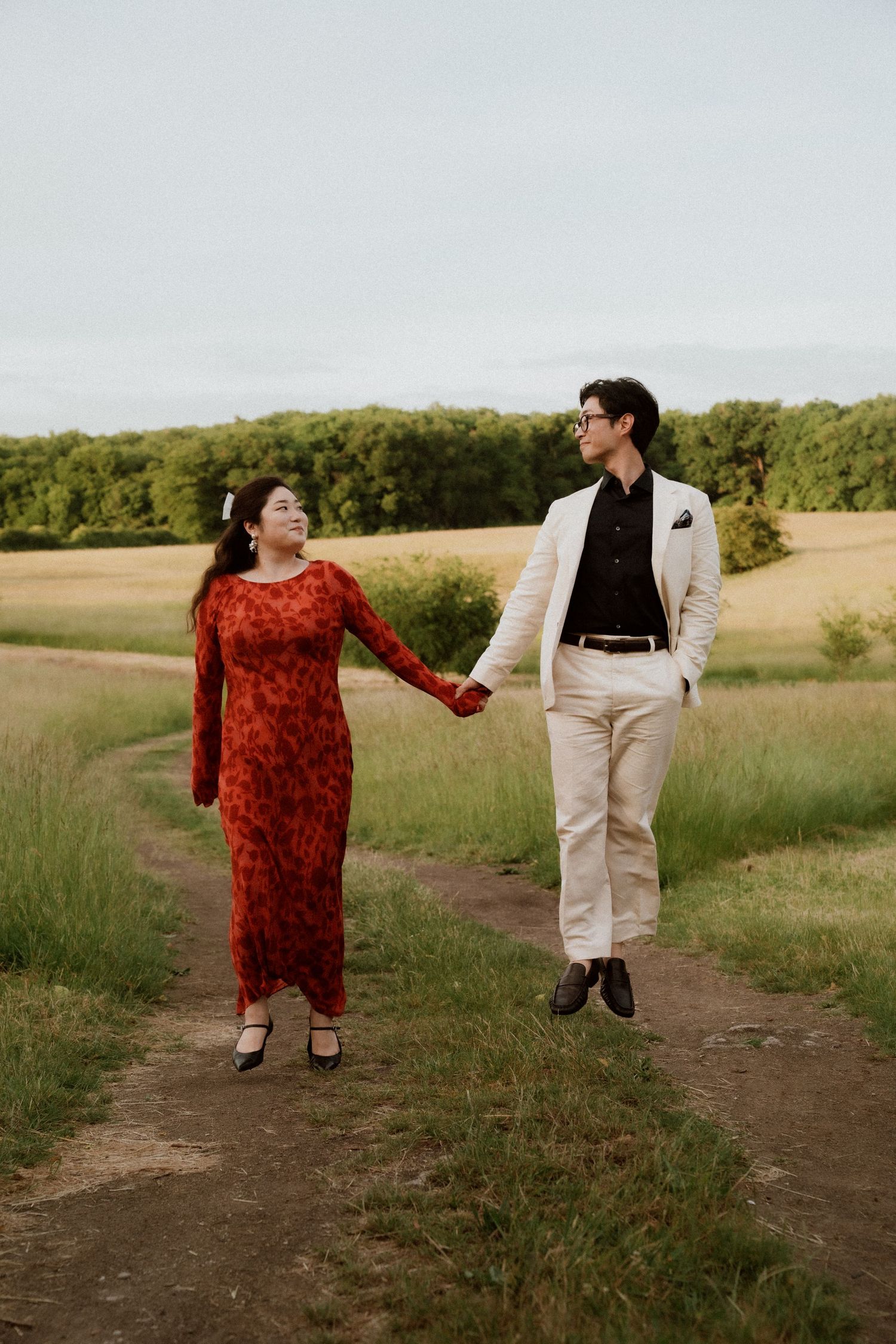 Couple in contrasting red dress and white suit walking together on a countryside path at sunset.