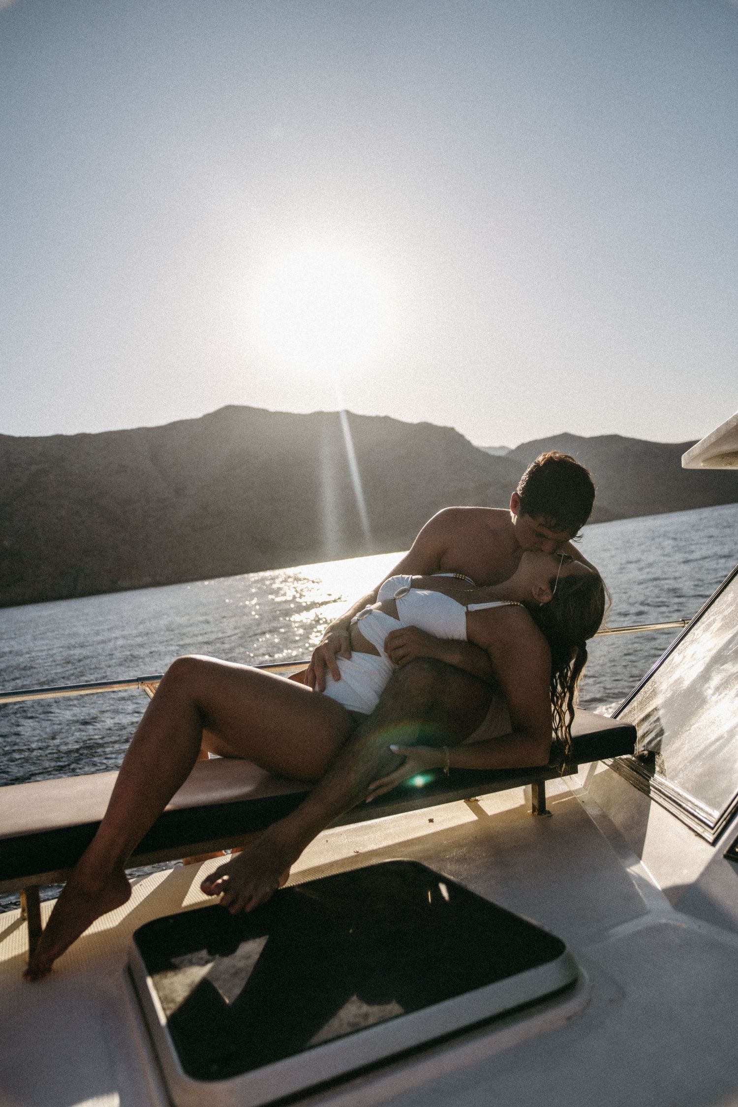 Intimate moment captured on a boat deck during sunset with mountains in the background. A woman lies across her husbands lap.