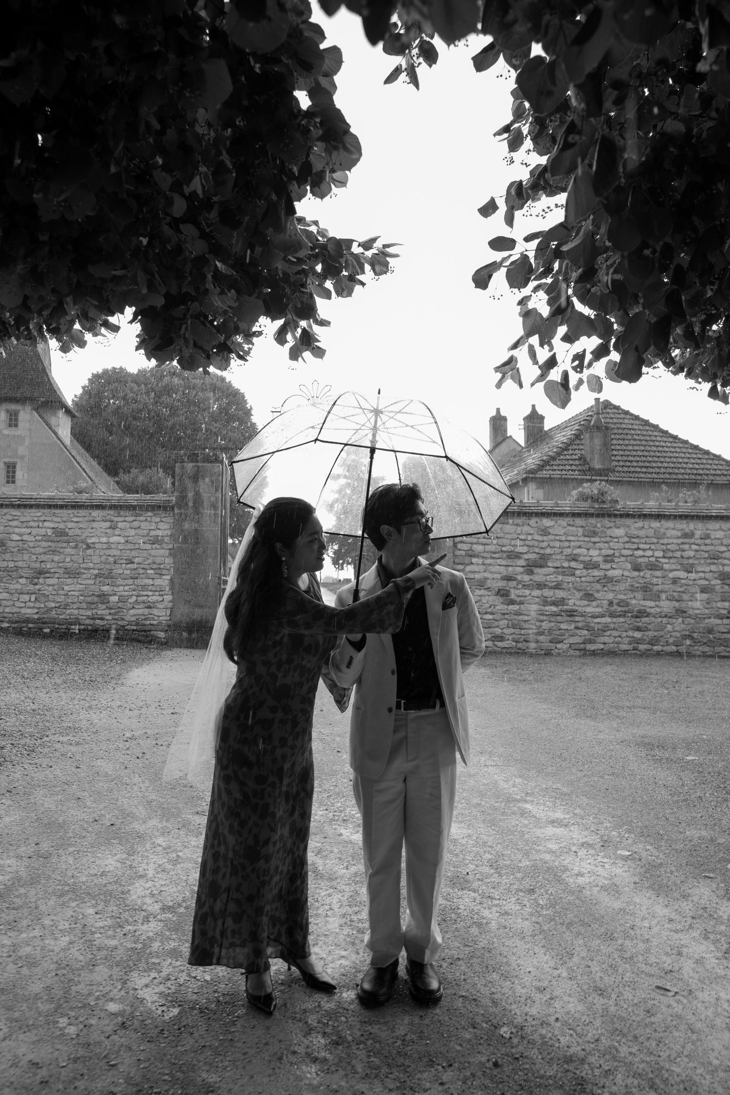 Vintage black and white photo of two figures with umbrellas standing beneath trees on a cloudy day.