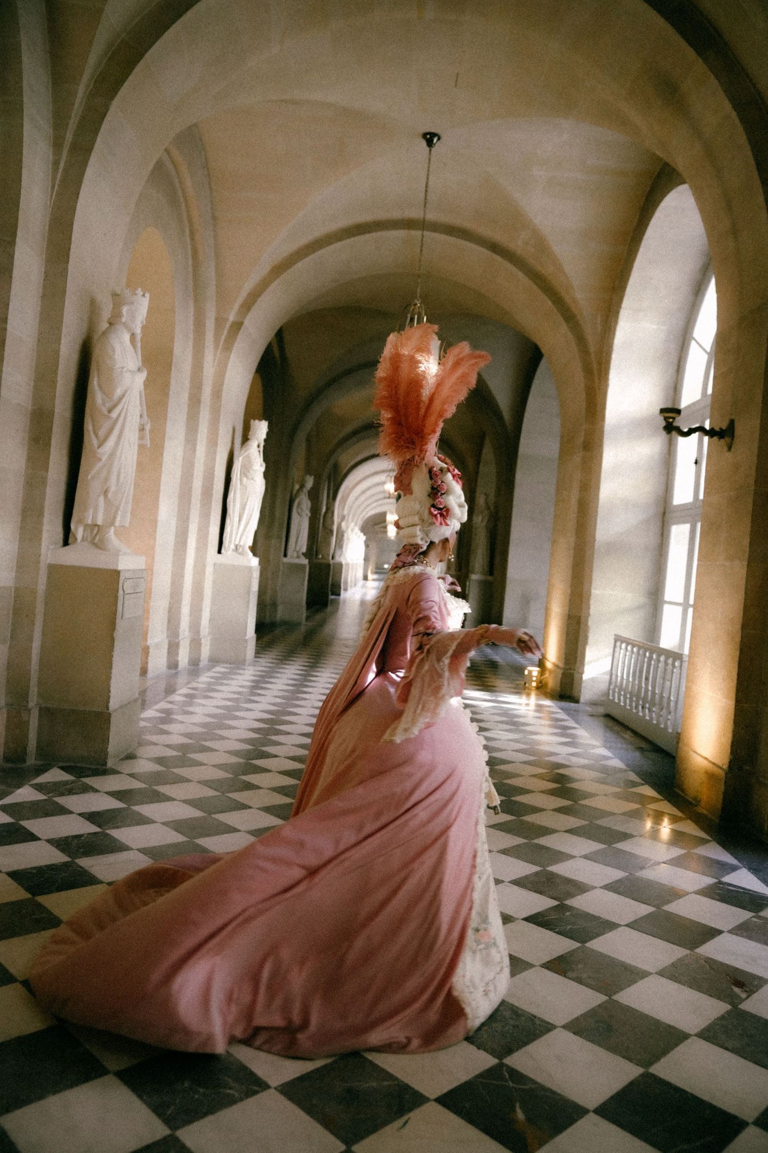 A flowing pink gown sweeps across a dramatic black and white checkered floor in a grand arched hallway in the Palace of Versailles