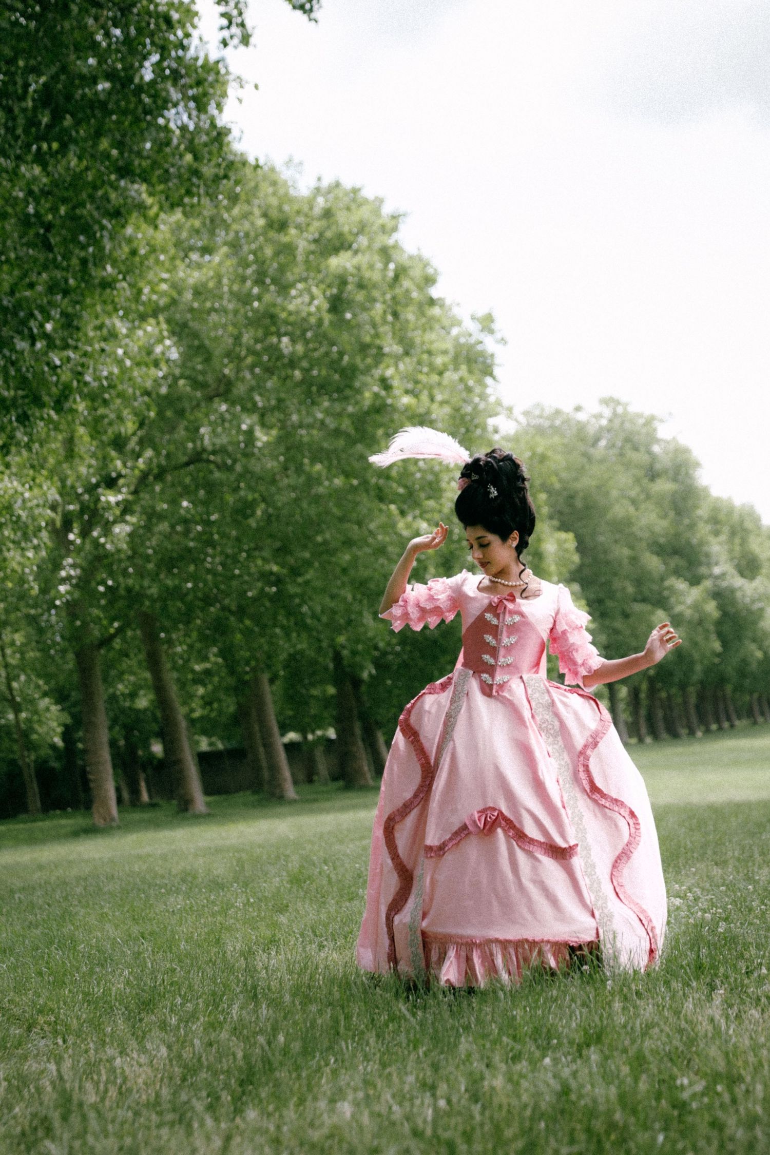 A person in a flowing pink vintage dress twirls gracefully in a serene park setting surrounded by green trees.