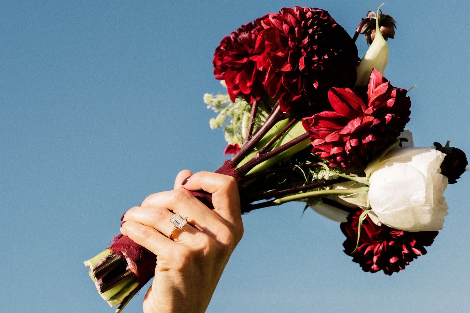 Hand holding a bridal bouquet of red dahlias and white flowers against a clear blue sky.