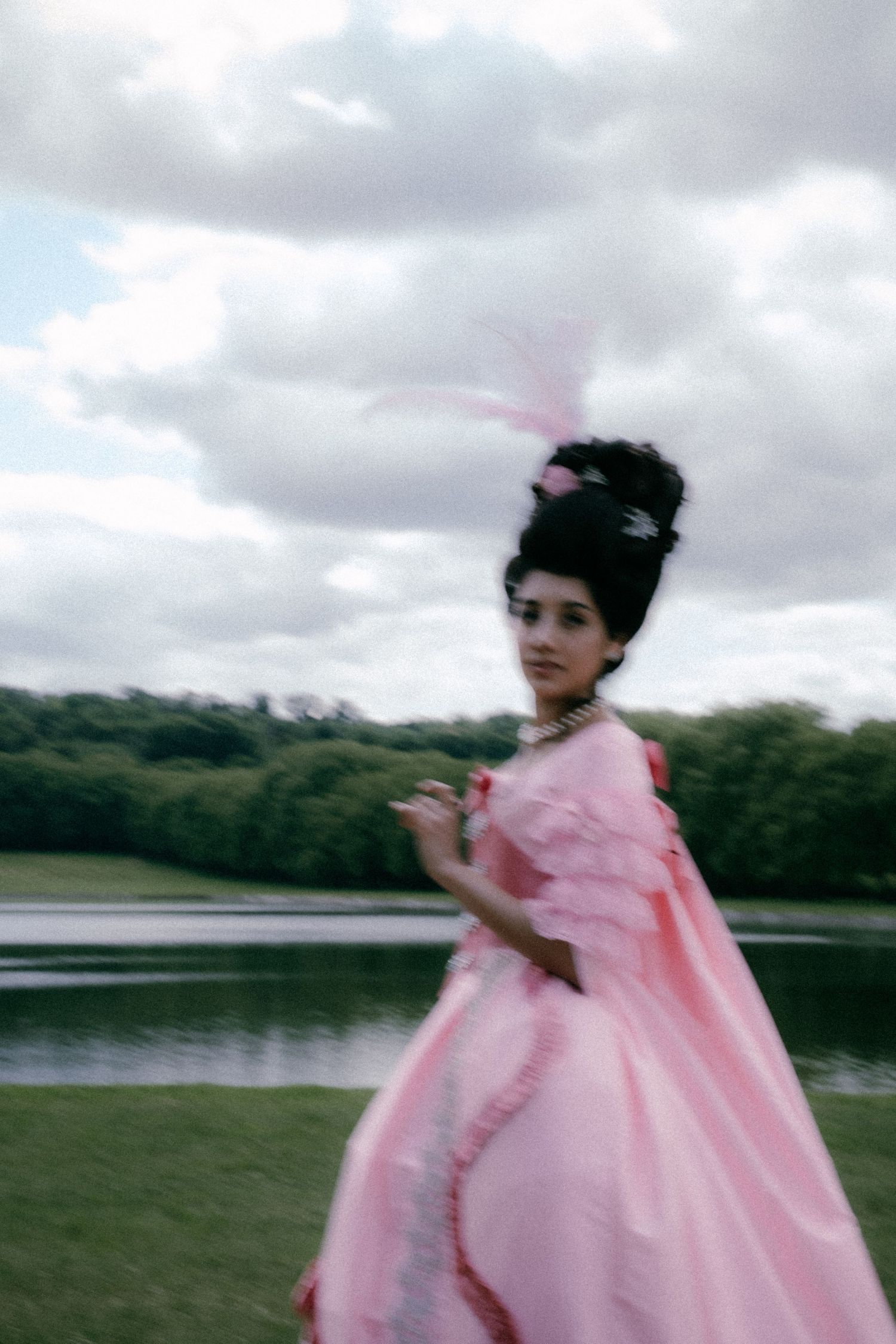 A figure in a flowing pink dress runs through a grassy field near a lake on an overcast day.