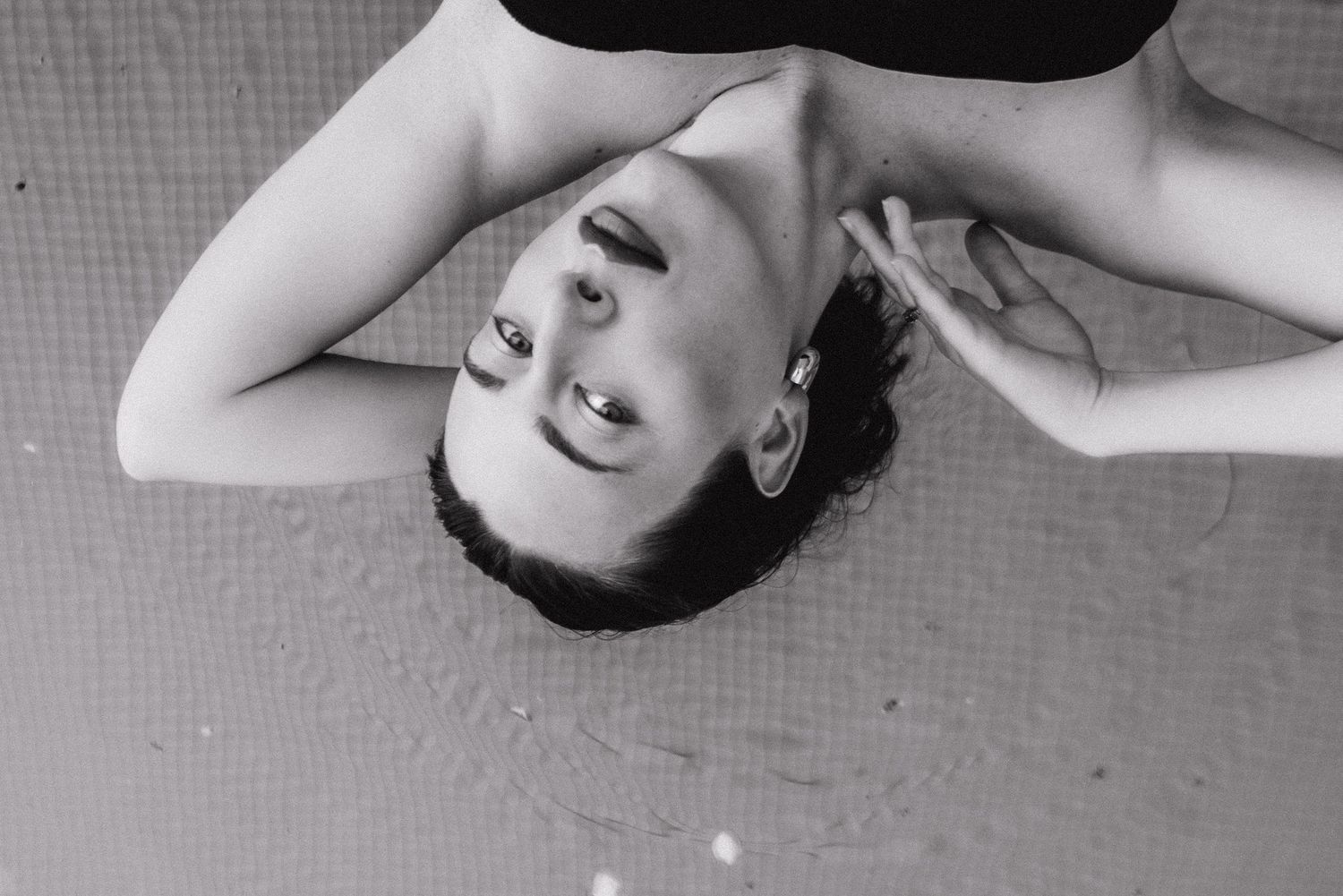 Black and white overhead photo of a young woman lying on the ground, smiling upward with wet hair.