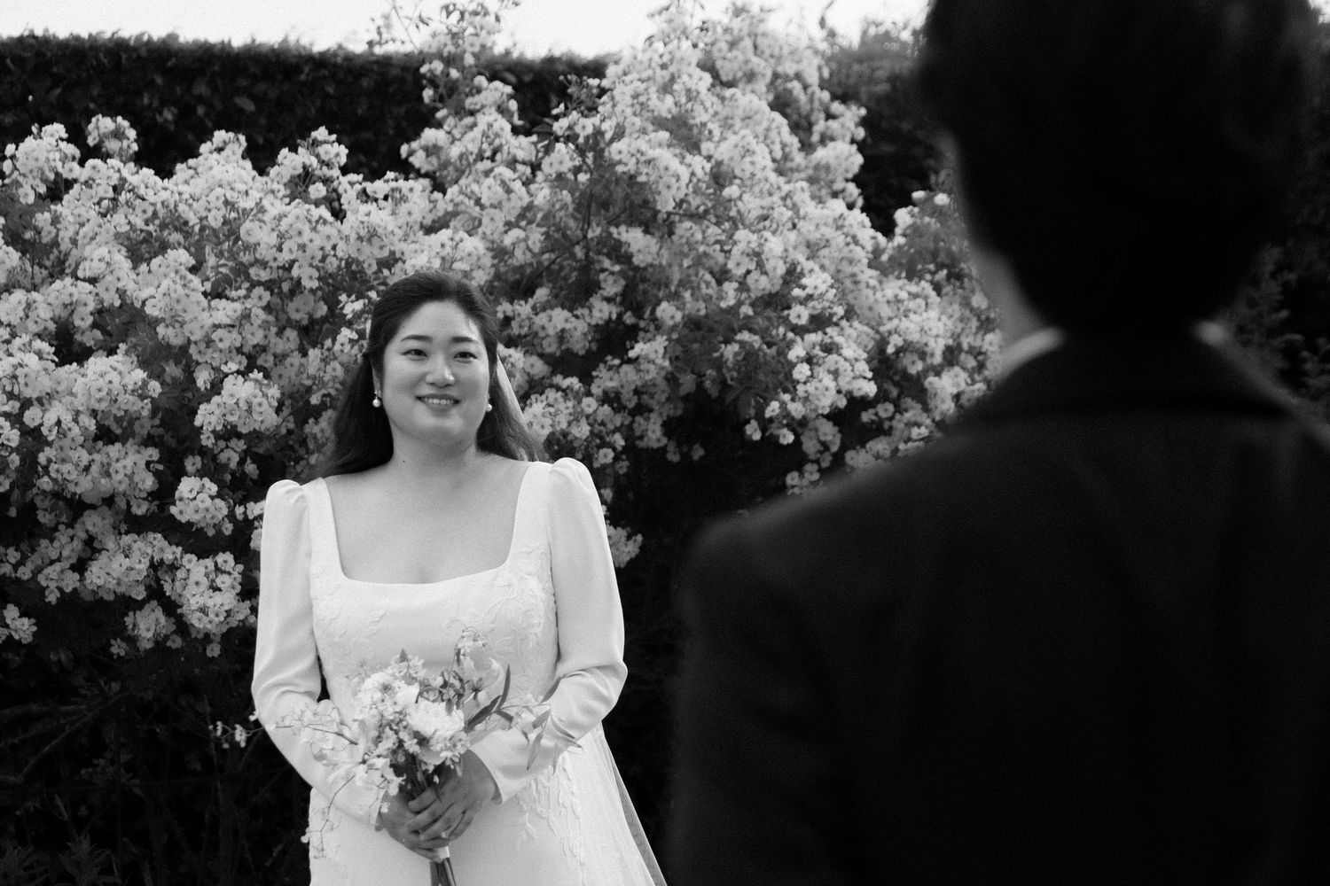 A bride in a white wedding dress poses outdoors with blooming white flowers and trees in the background.