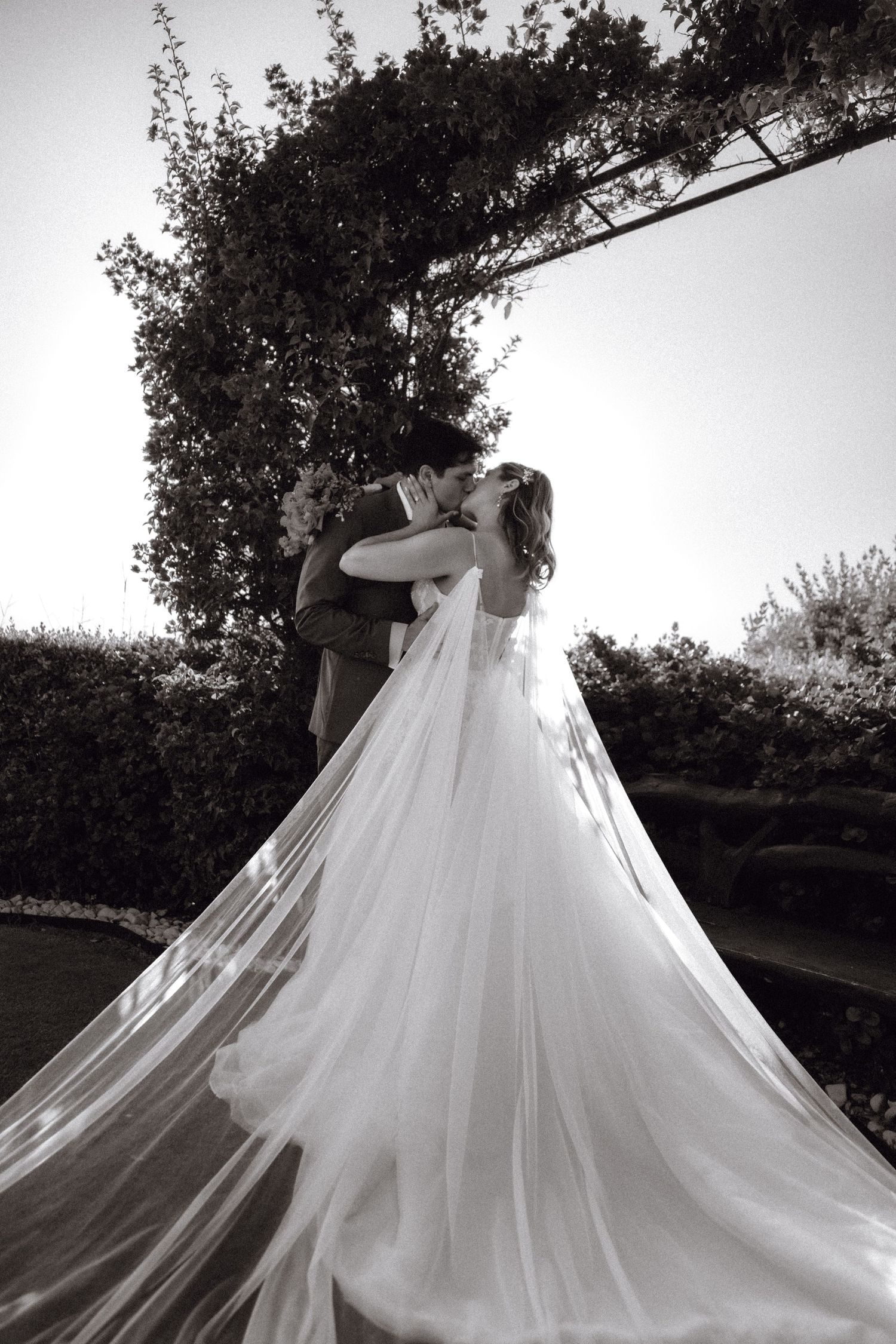 Flowing white wedding dress train billows dramatically against a garden backdrop in black and white photography.