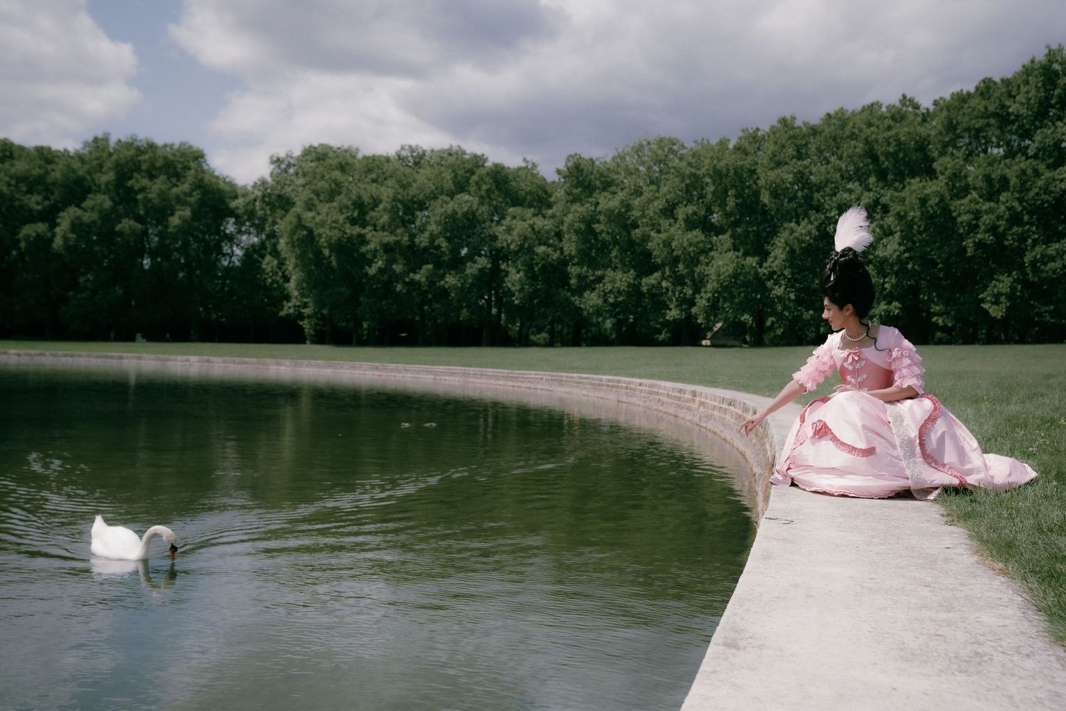 A person in a pink dress stands on a curved pathway by a serene pond with a white swan swimming nearby.