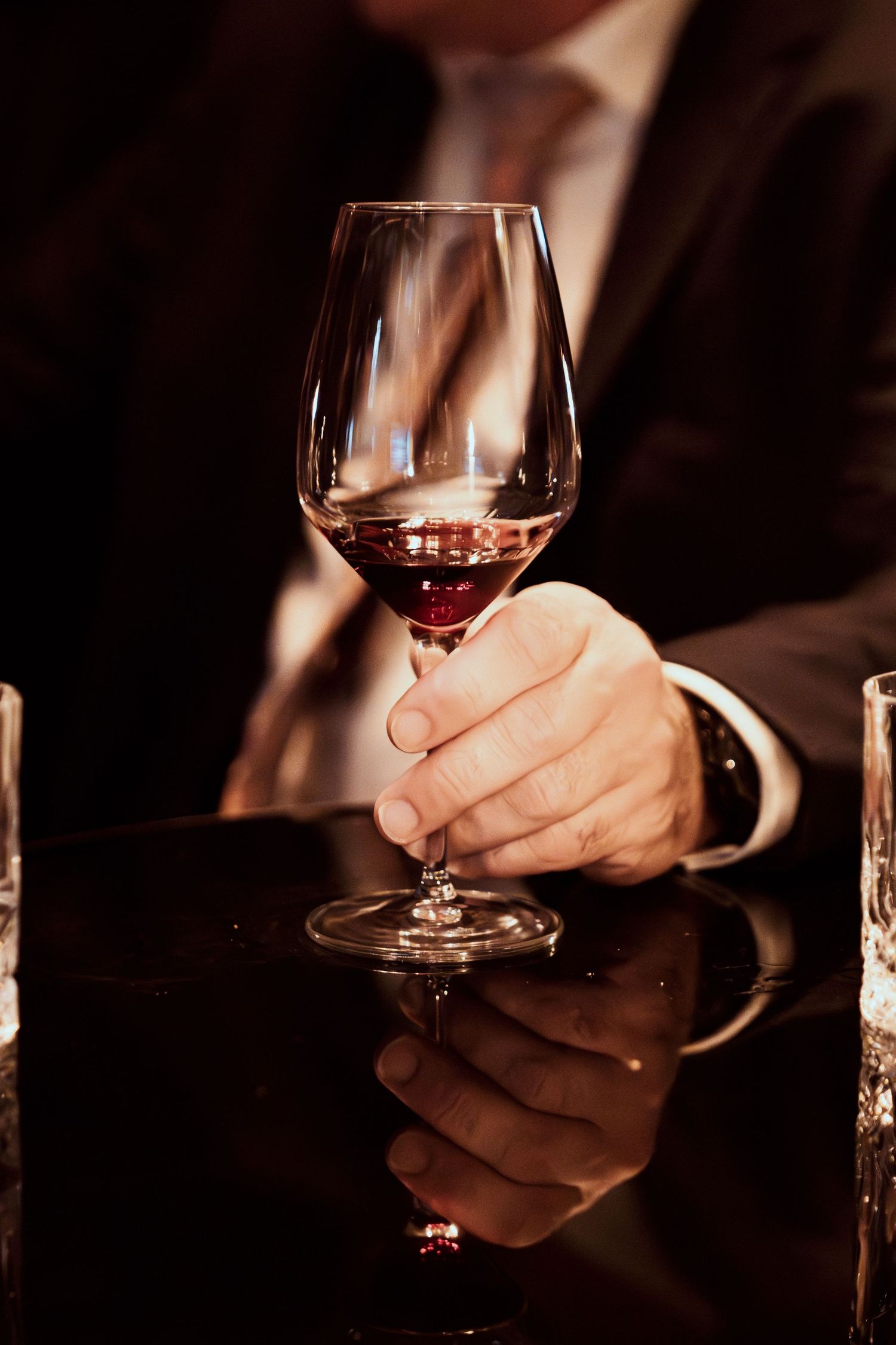 Hand in formal attire holds a glass of red wine against dark reflective surface in moody lighting.