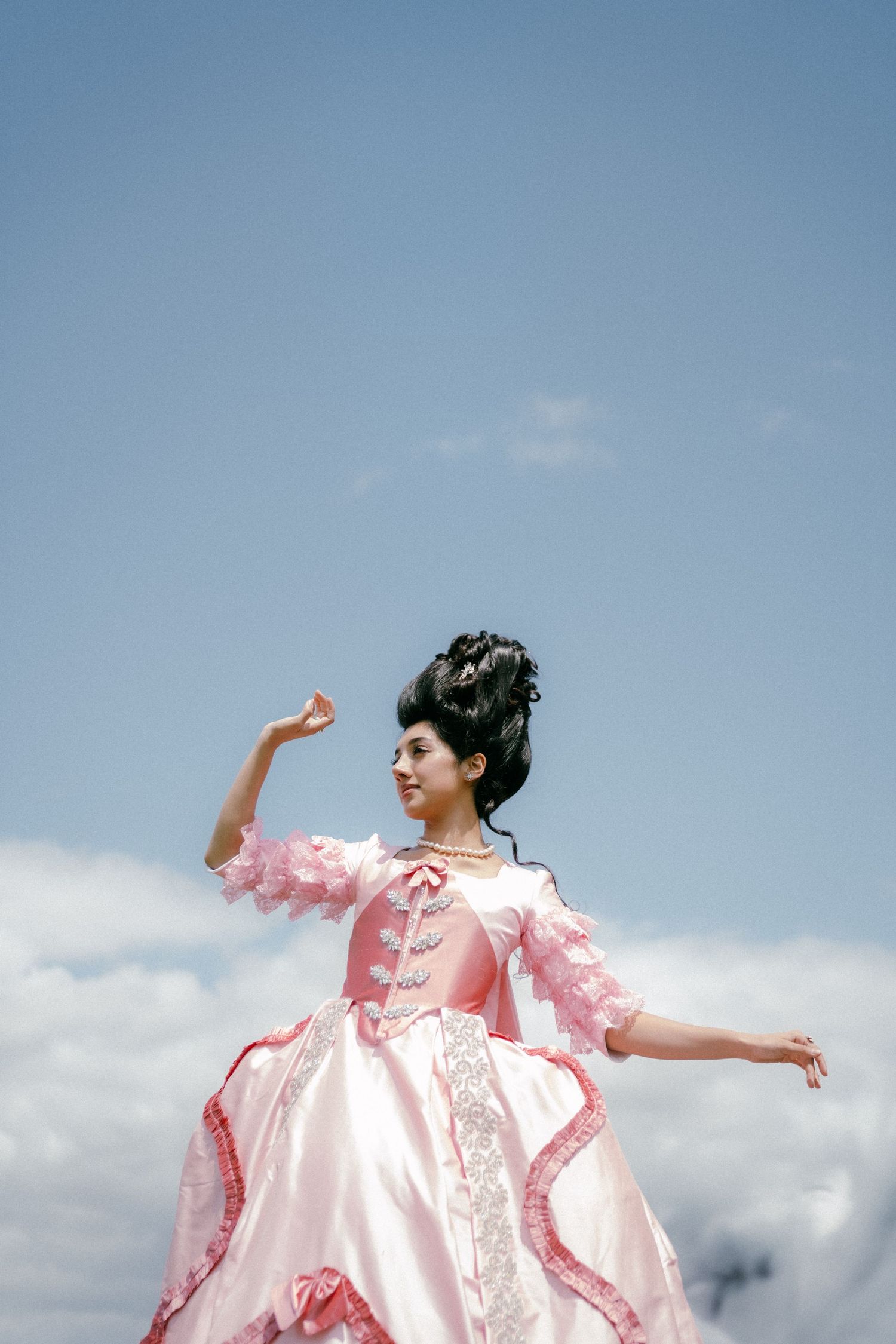 A figure in a flowing pink dress with ruffled details twirls gracefully against a cloudy blue sky backdrop.