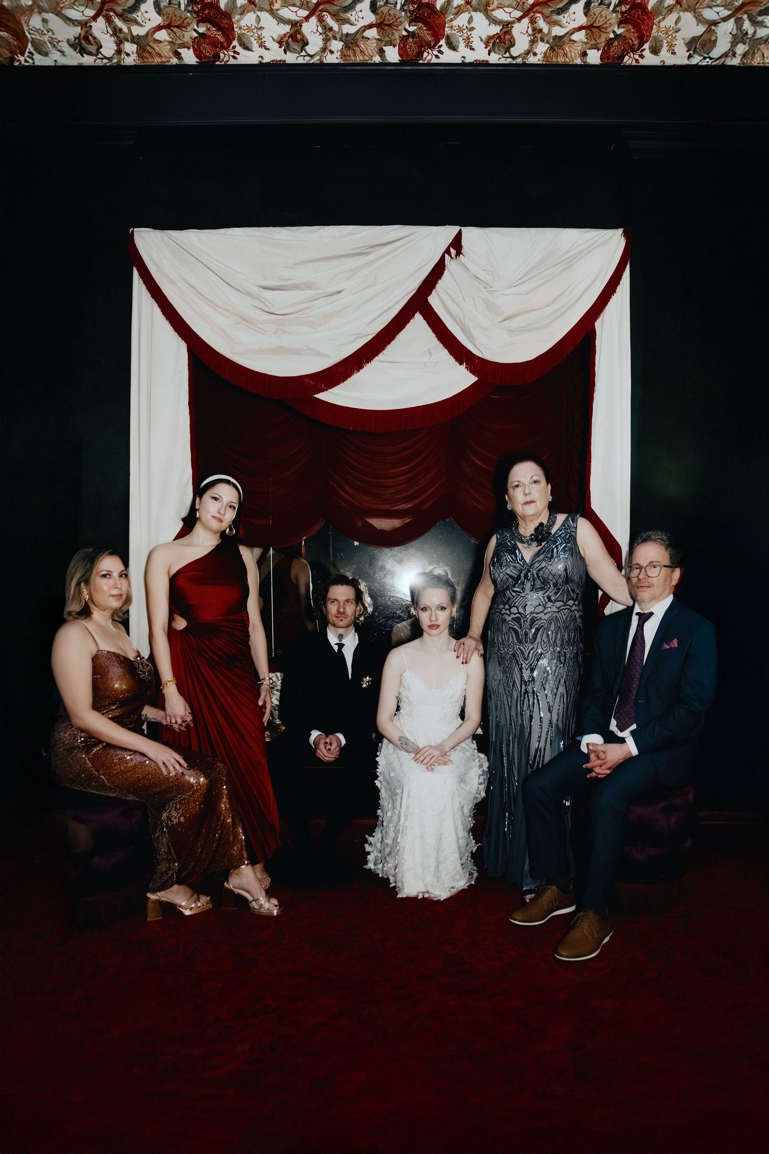 Wedding party gathering in front of white and burgundy draped backdrop in dark room.