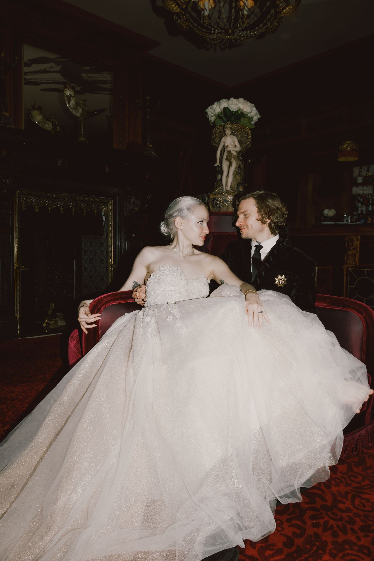 A flowing white wedding dress billows across a deep red carpet in a dimly lit ballroom.