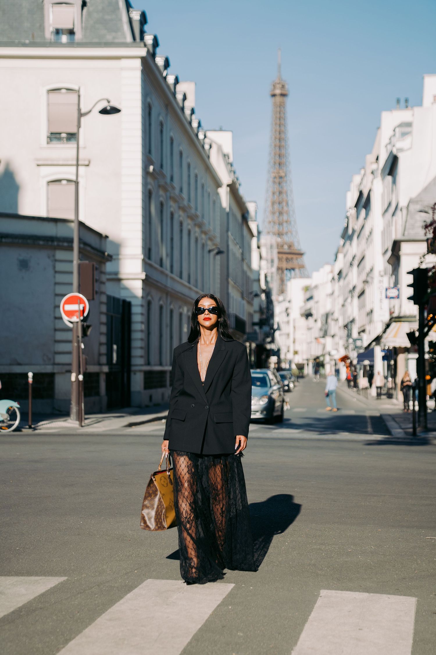 Stylish figure in black attire walking on a Parisian street with the Eiffel Tower visible in the distance.