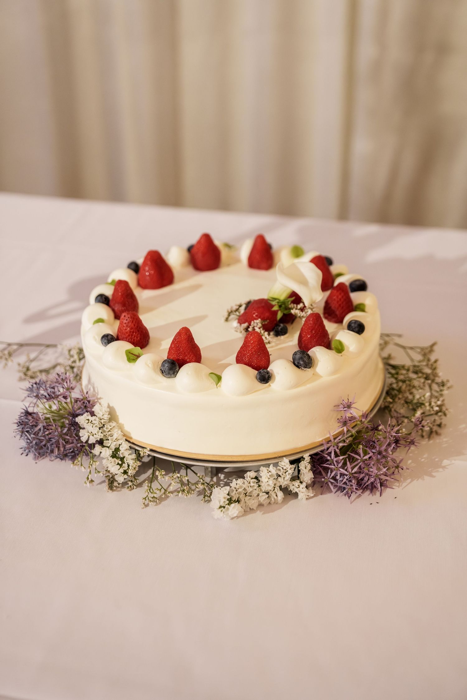 White pavlova dessert decorated with fresh strawberries and blueberries on a white surface with dried flowers.
