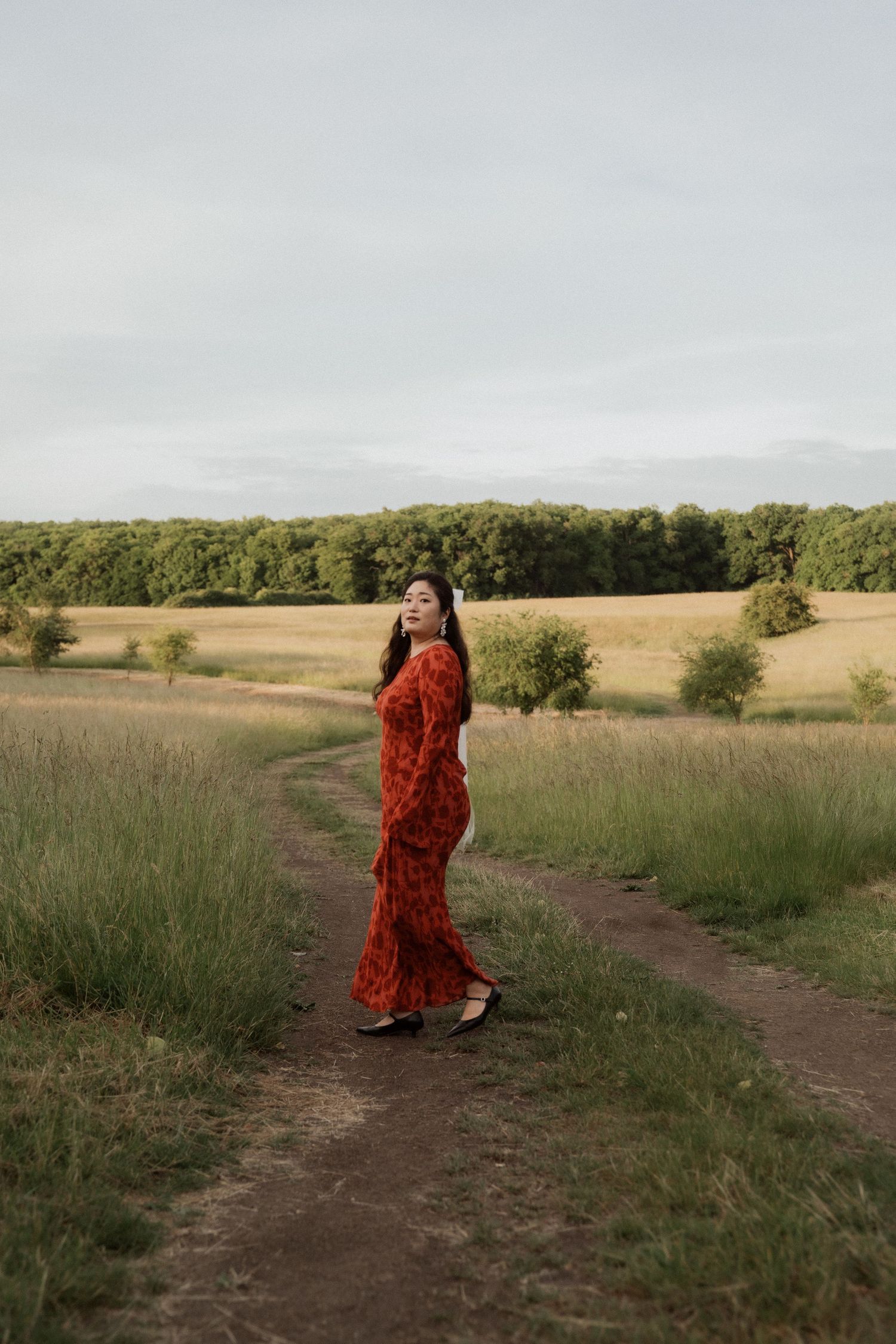 Person in a flowing red dress walking through a serene rural landscape with dirt paths and open fields.