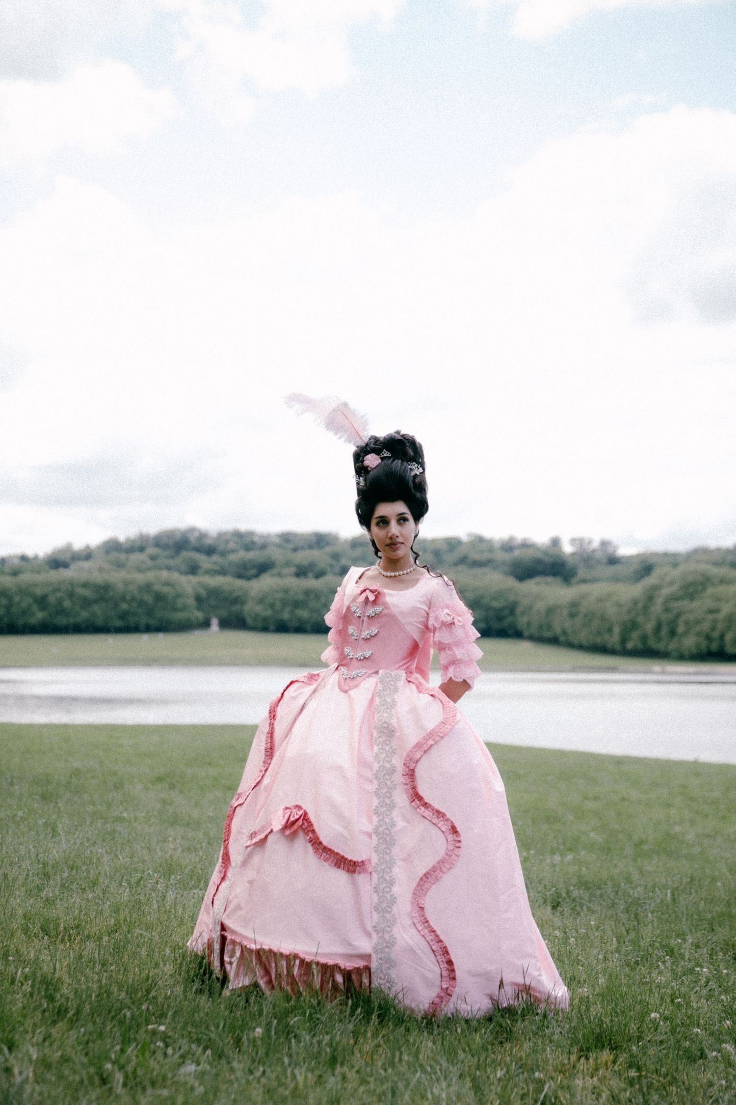 A person in a flowing pink ruffled dress runs through a serene meadow landscape beside a lake.