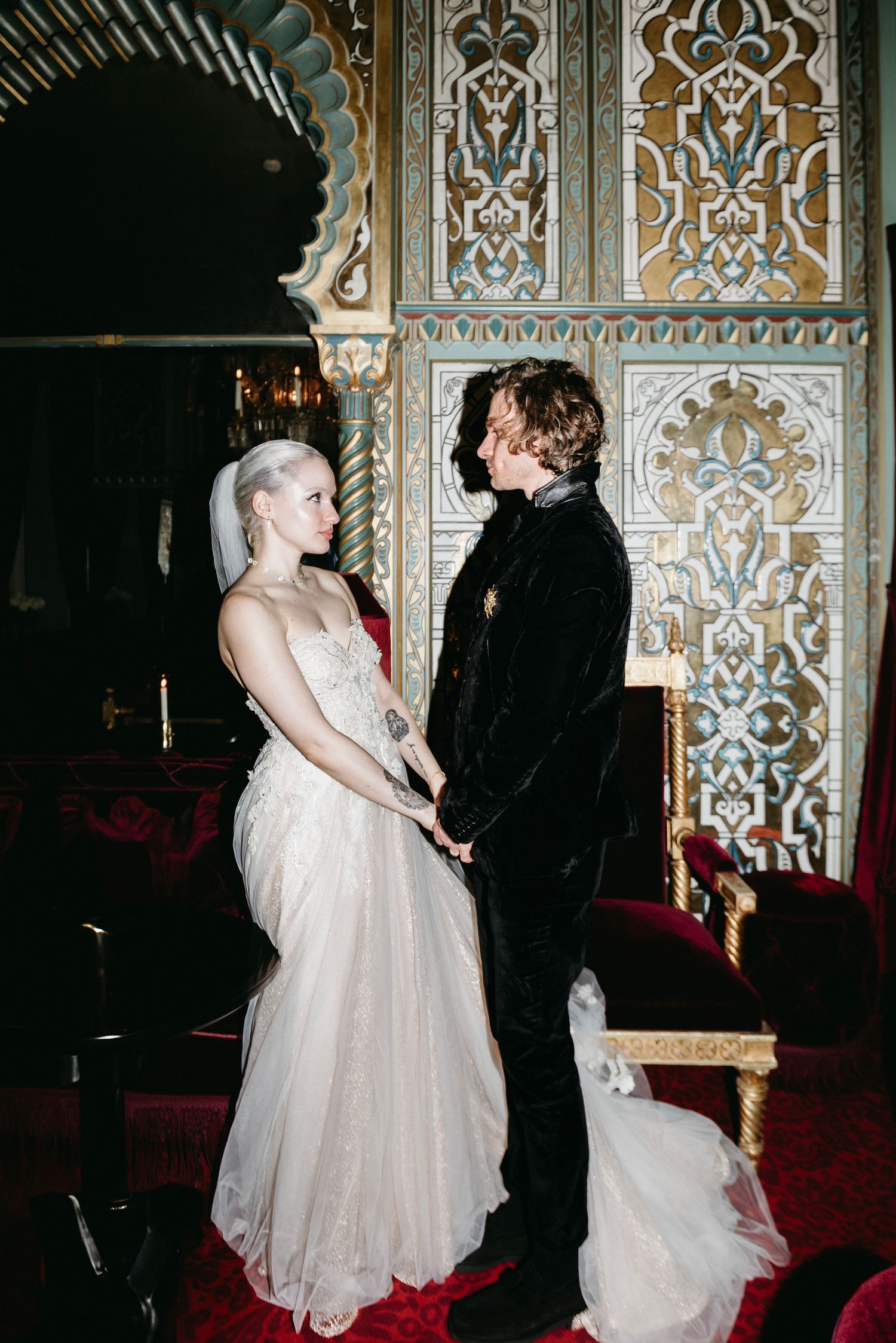Couple in formal wedding attire standing against ornate metalwork wall with intricate patterns.