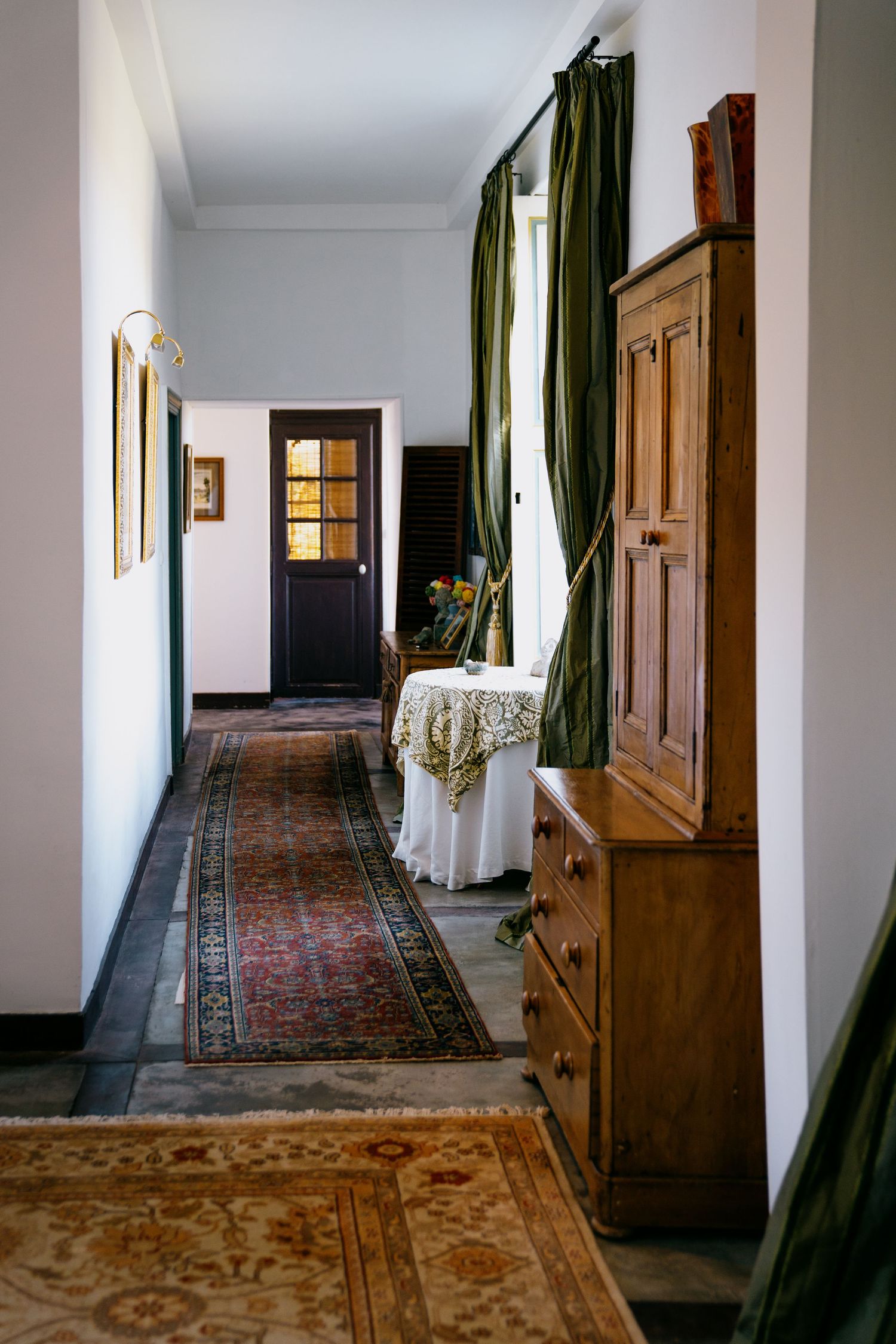Long hallway with wooden furniture and green curtains features an ornate patterned carpet runner.