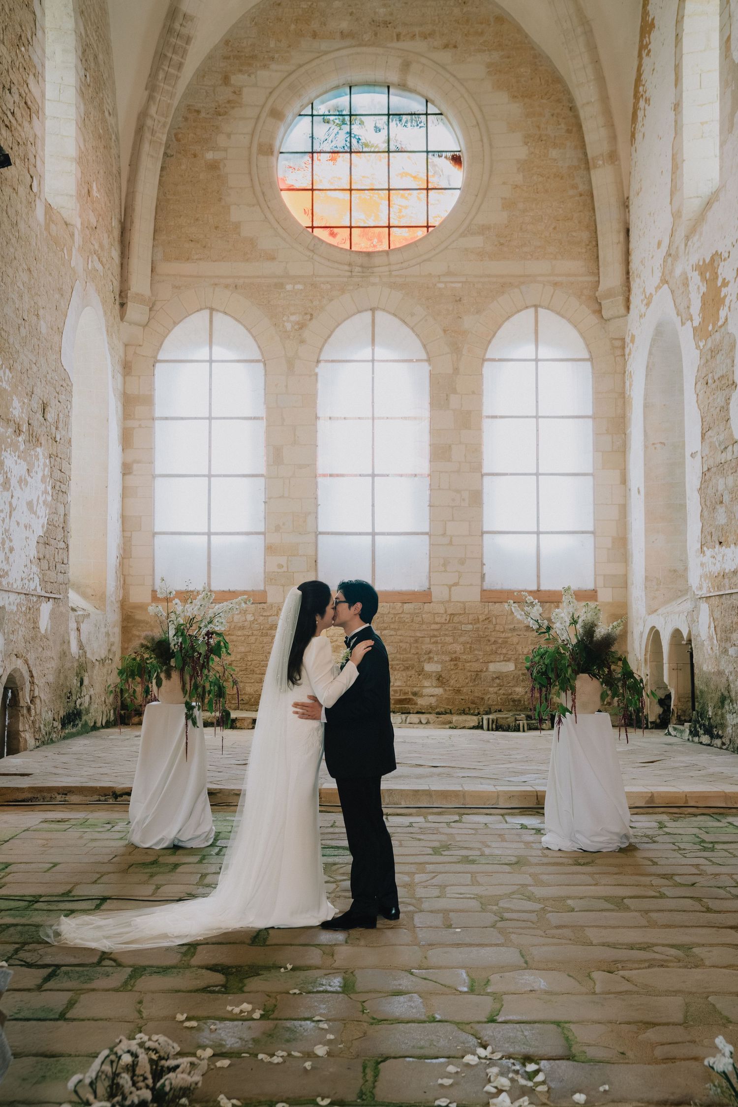 A romantic wedding photoshoot in a historic stone chapel with arched windows and scattered rose petals.