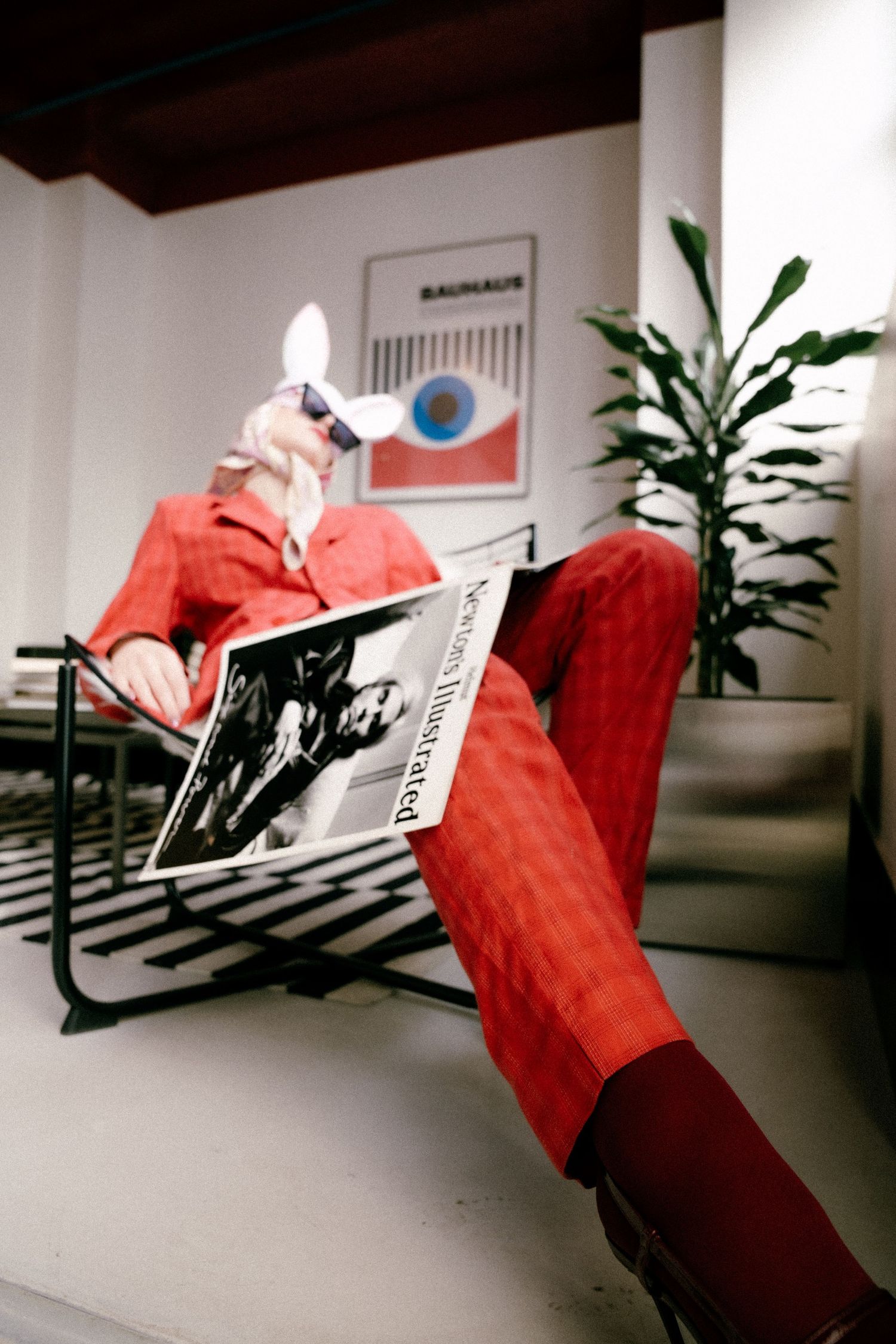 Paris fashion editorial with retro styling. Model in red with sunglasses and bunny mask, reading a magazine in curated interior space.