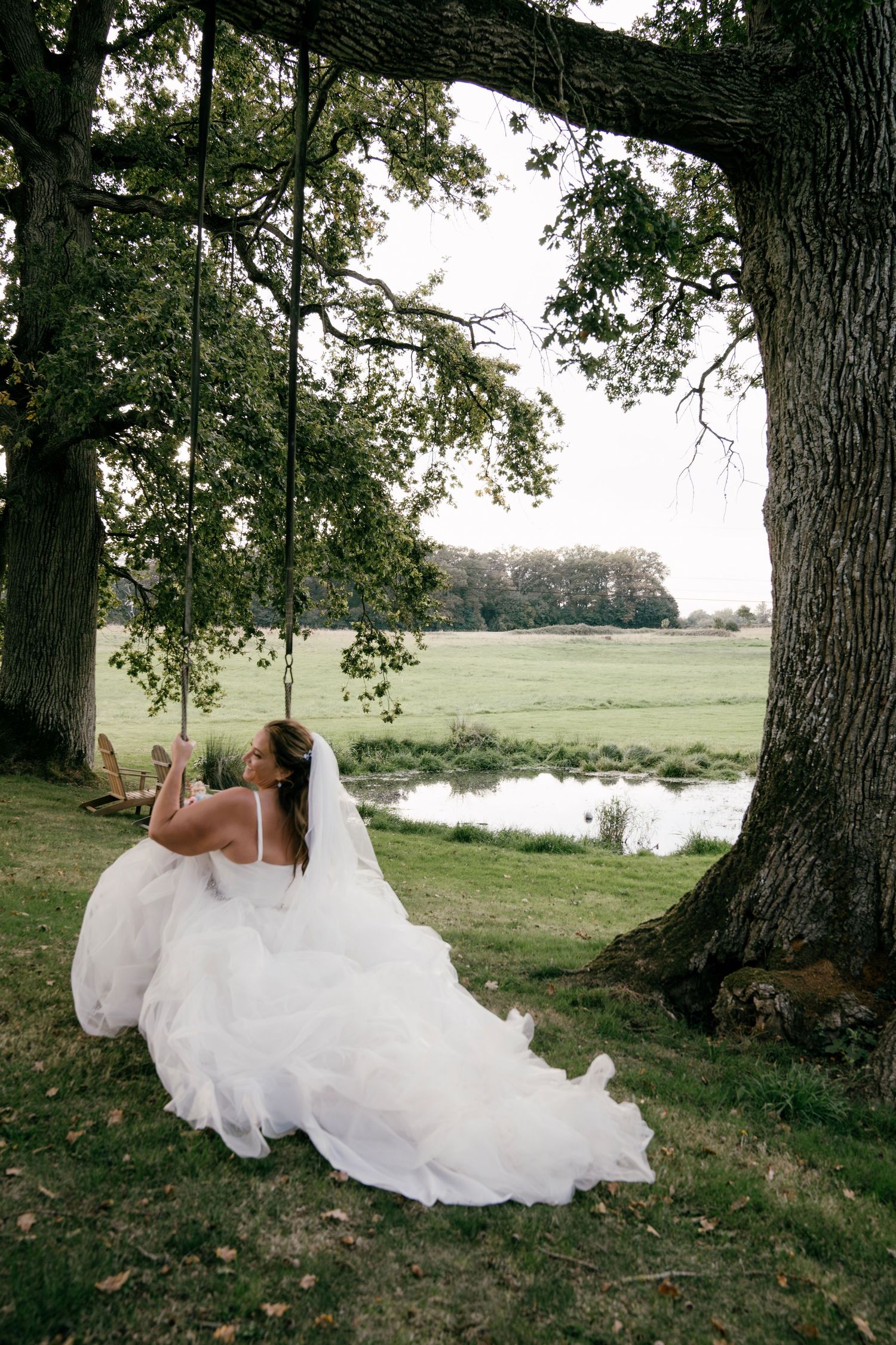 Bride in white gown sitting on a tree swing in a lush green countryside setting with a pond in the background.