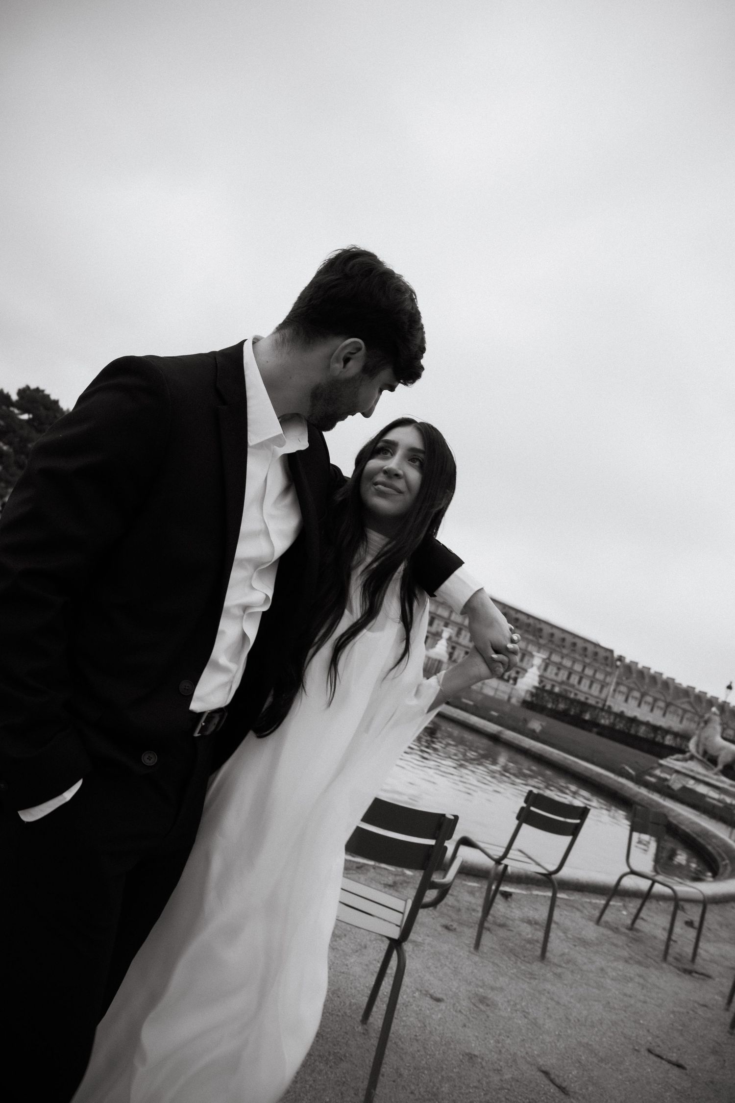 A couple shares an intimate black and white moment outdoors with chairs and a building in the background.