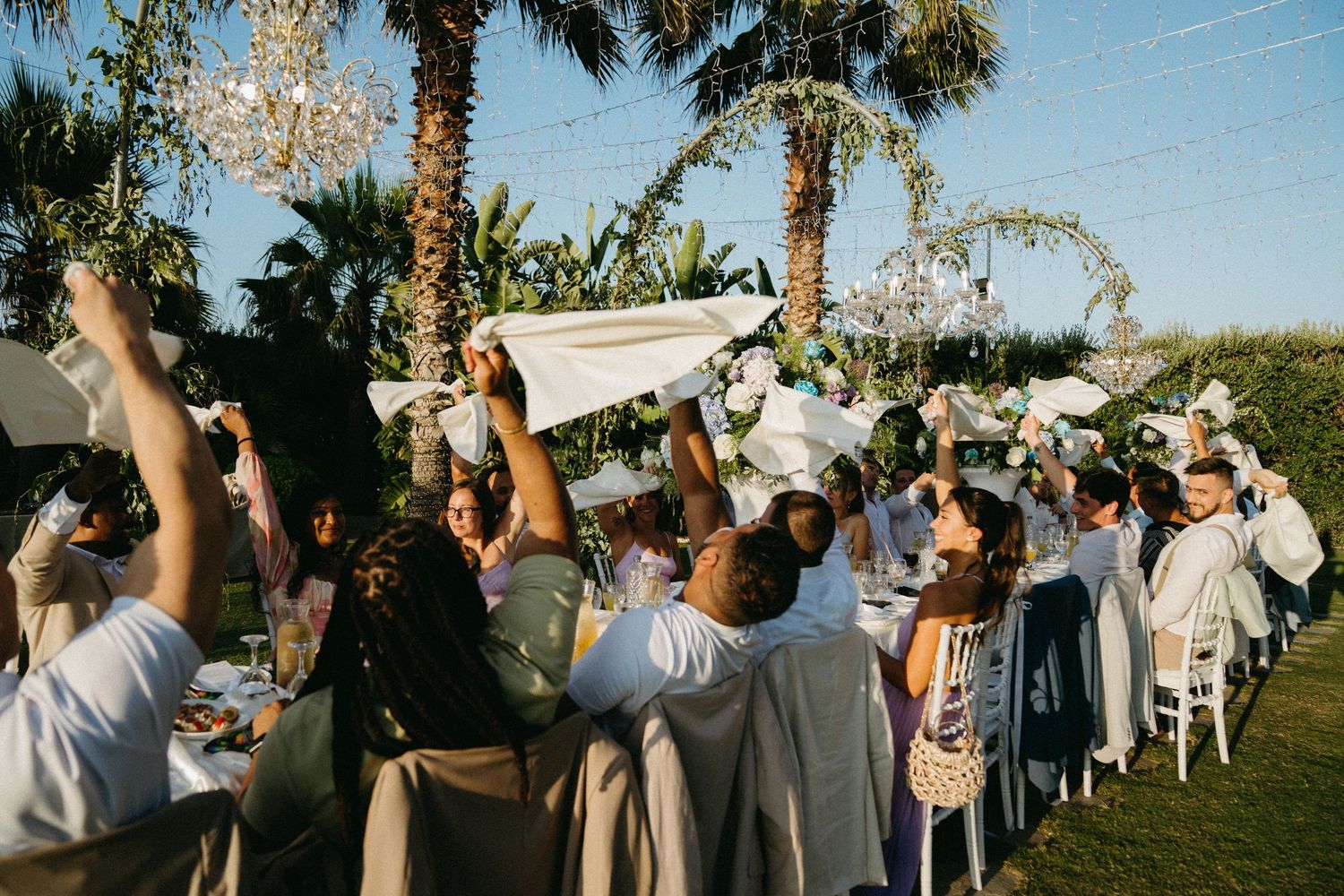 Wedding guests stand outdoors under palm trees watching a solar eclipse through protective glasses at a daytime ceremony.