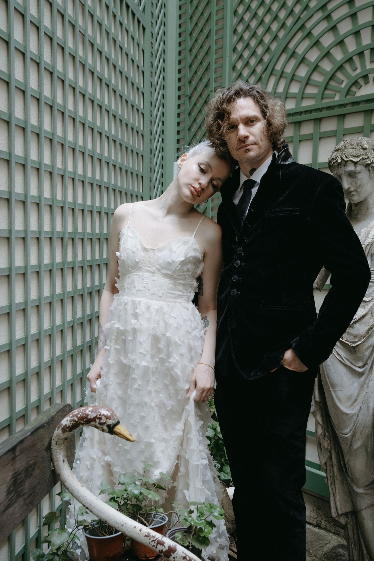 Wedding couple poses against a green lattice wall with classical statues in an elegant garden setting.