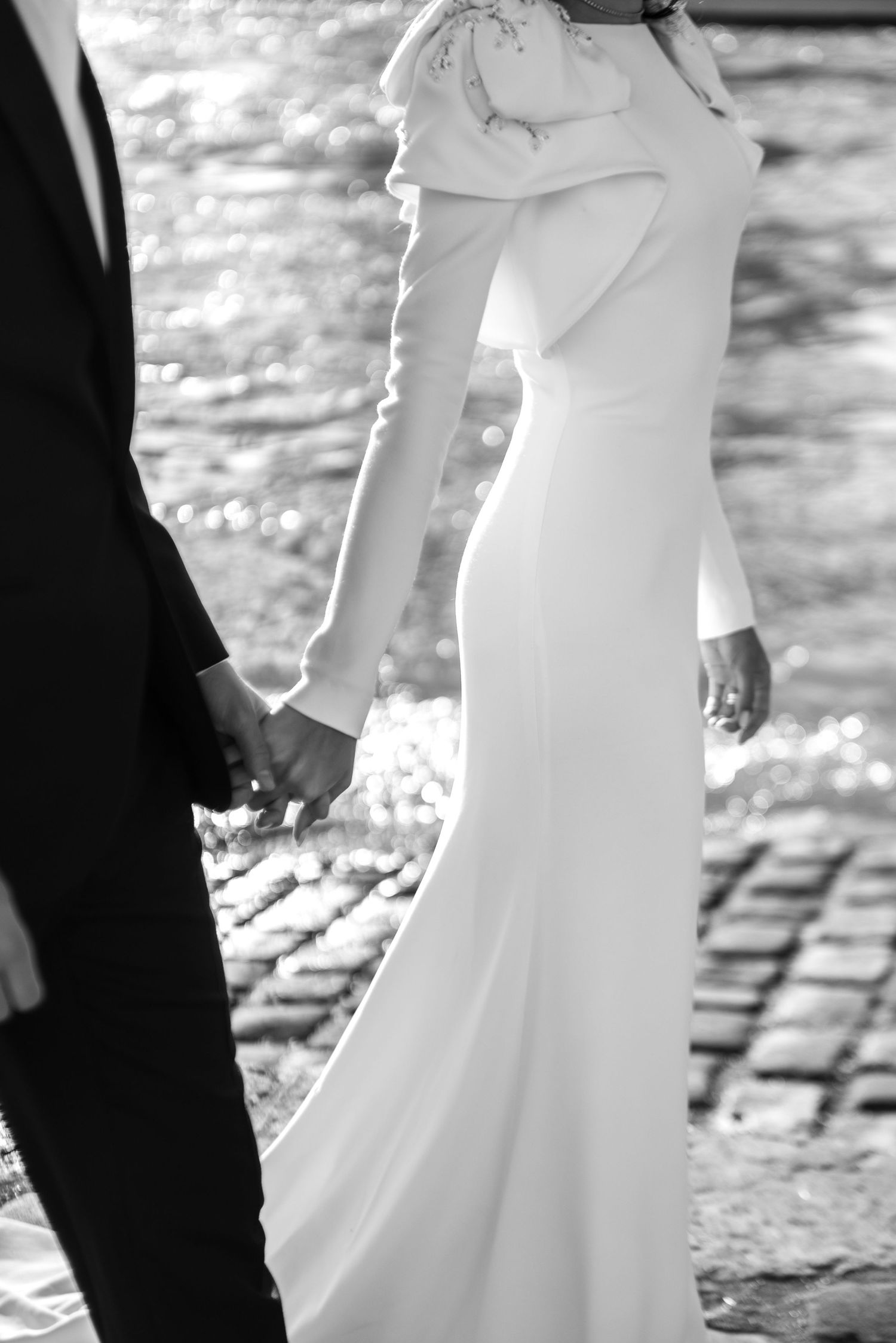 Black and white shot of couple holding hands by the Seine, bride in long-sleeved gown, cinematic Paris wedding editorial.