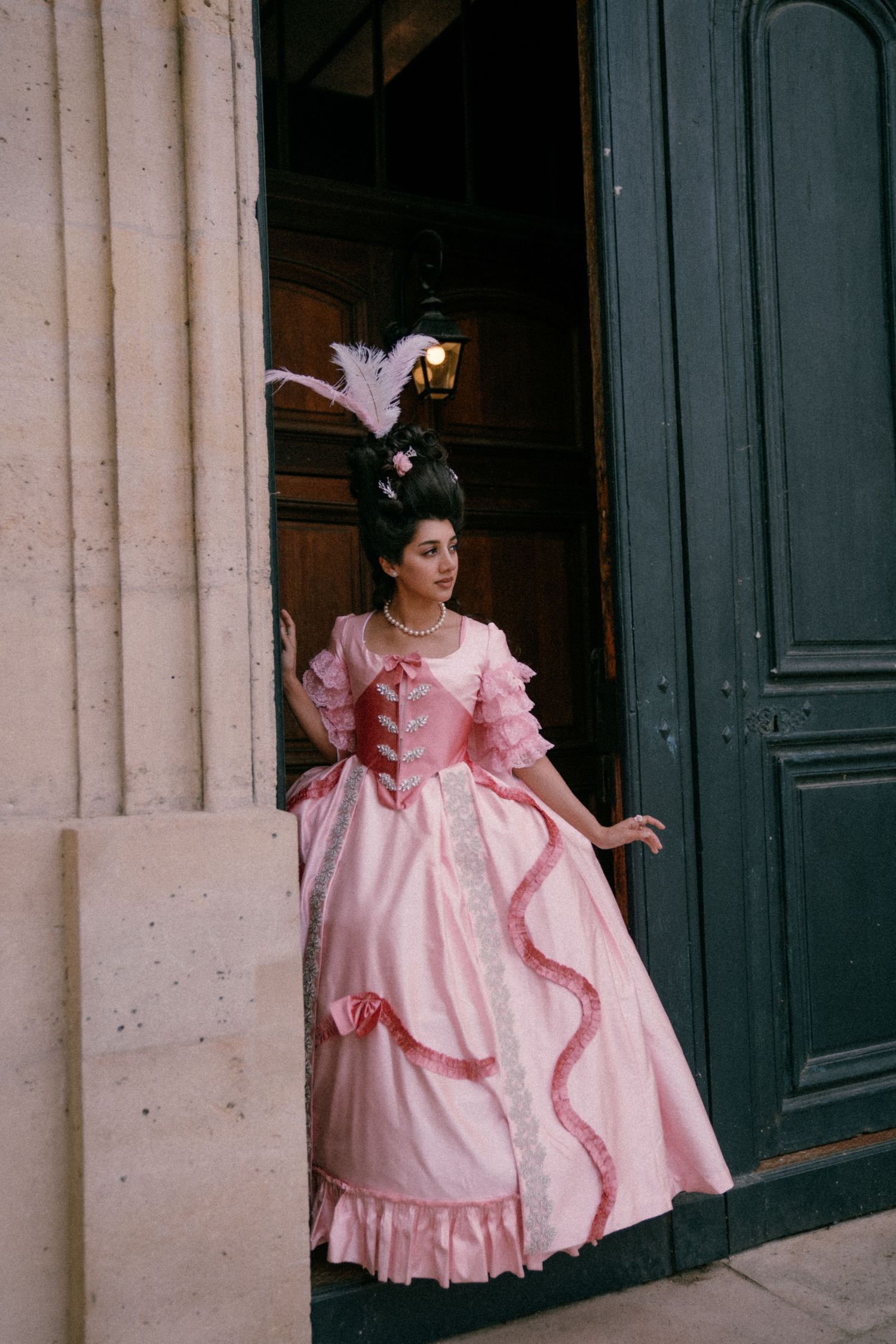 A person in a pink vintage dress with ruffles poses in front of dark blue doors against a stone building.