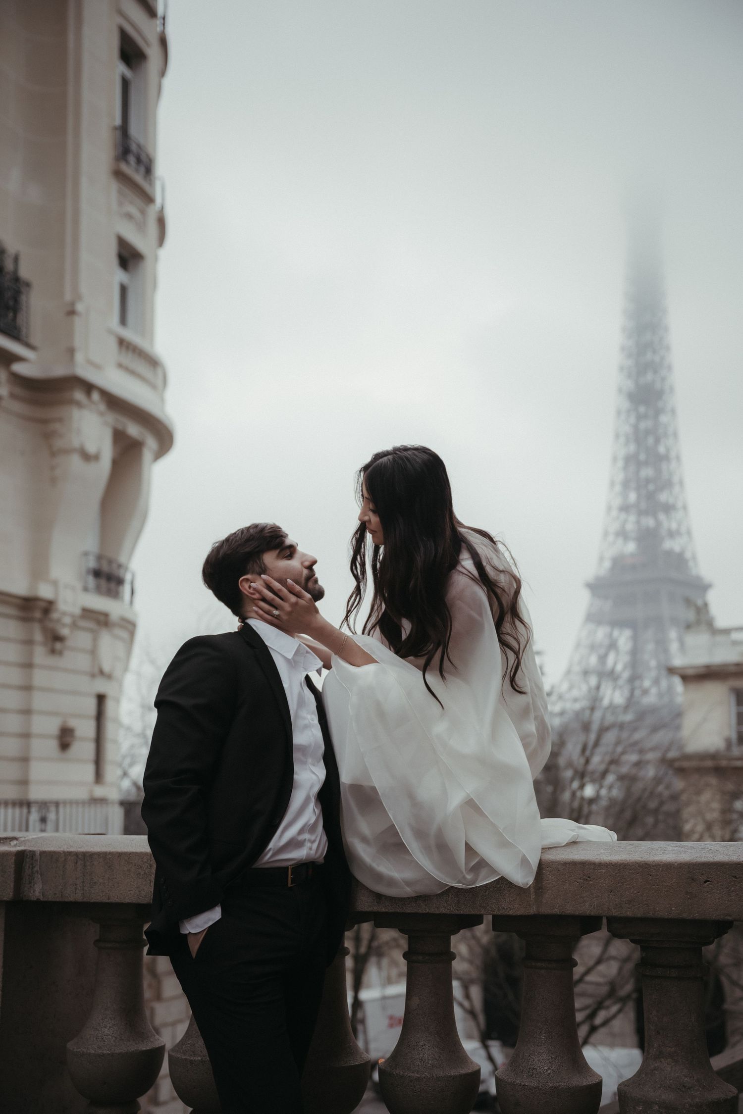 A romantic couple shares an intimate moment on a balcony with the iconic Eiffel Tower visible through fog in Paris.
