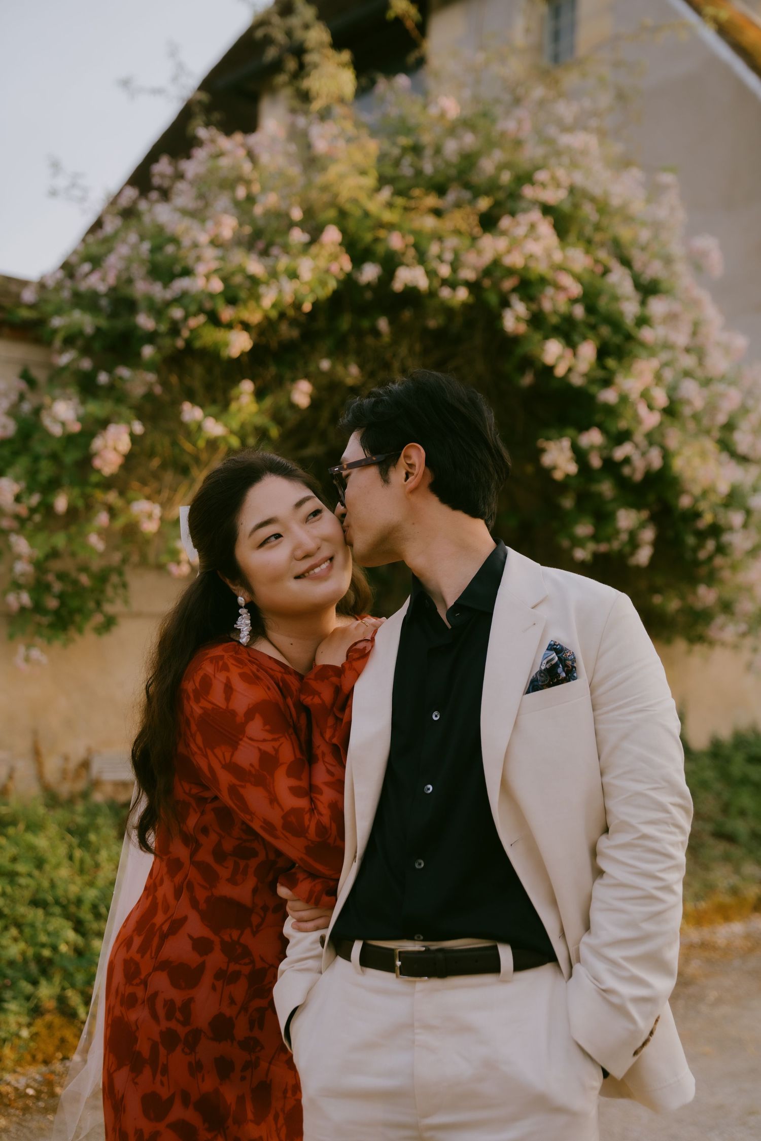 A couple shares romantic moments in front of a blooming rose garden wall wearing red and white outfits.