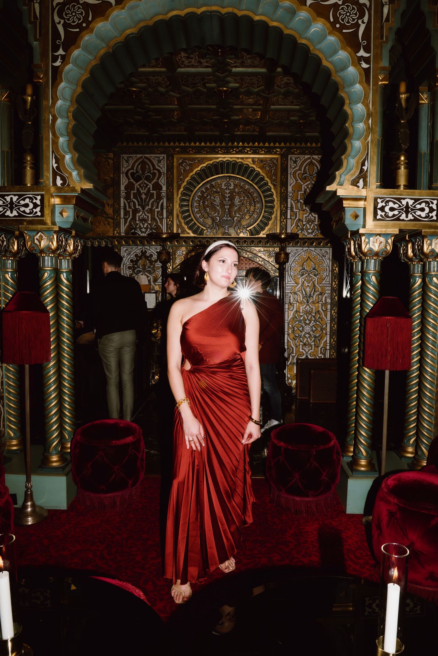 Elegant figure in a flowing red gown poses in an ornate room with Moorish architecture and velvet seating.