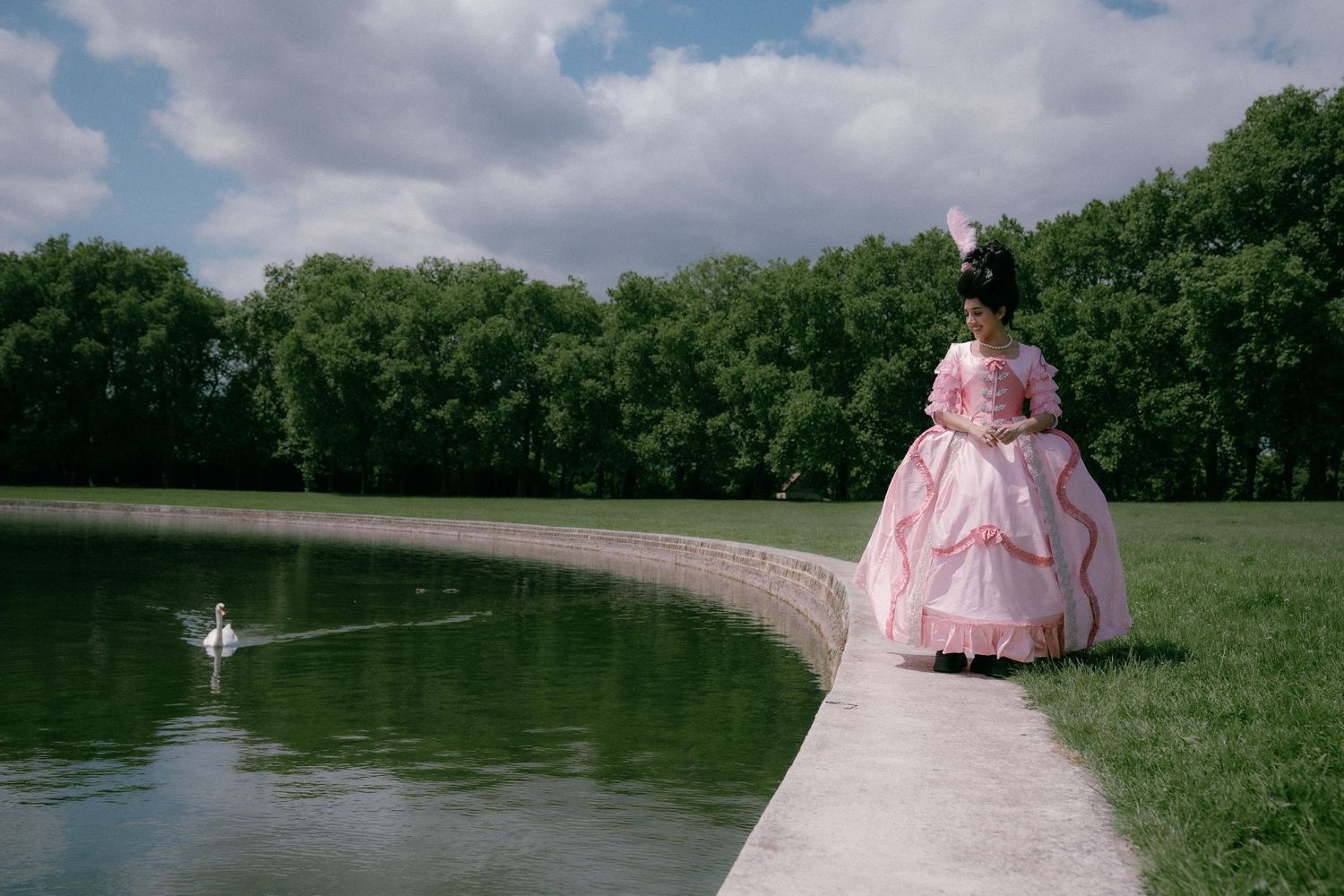 A person in a pink Victorian-style dress stands near a serene lake surrounded by lush green trees and cloudy skies.
