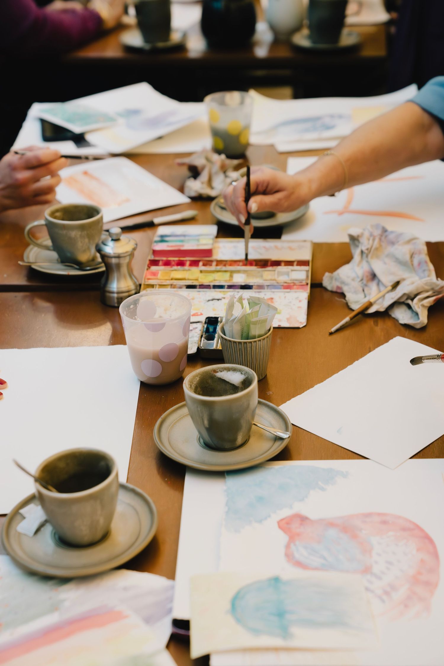Coffee cups and watercolor paintings scattered across a wooden table during an art session.