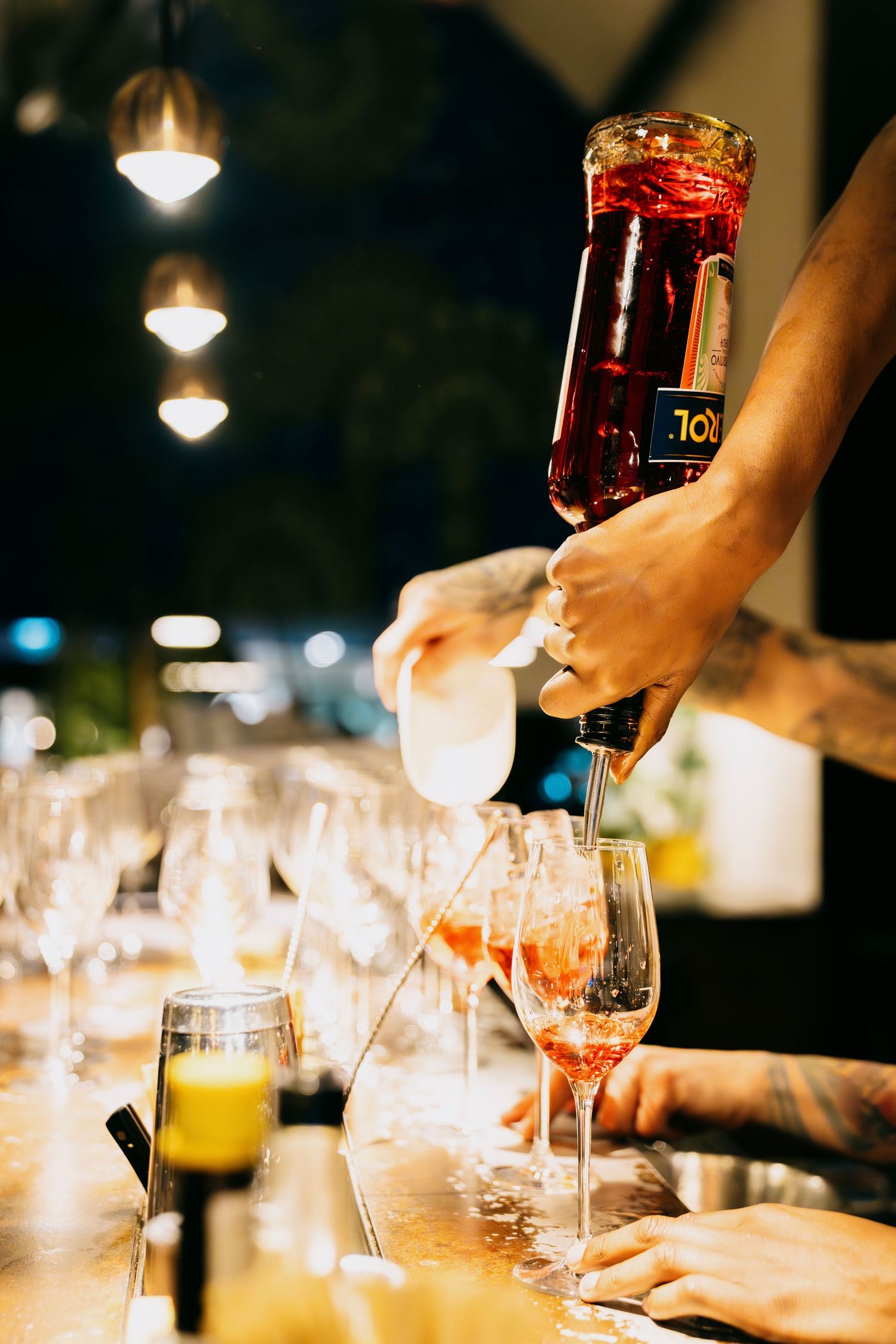 Hands pouring drinks and serving beverages at a dimly lit bar or restaurant with wine glasses on the table.