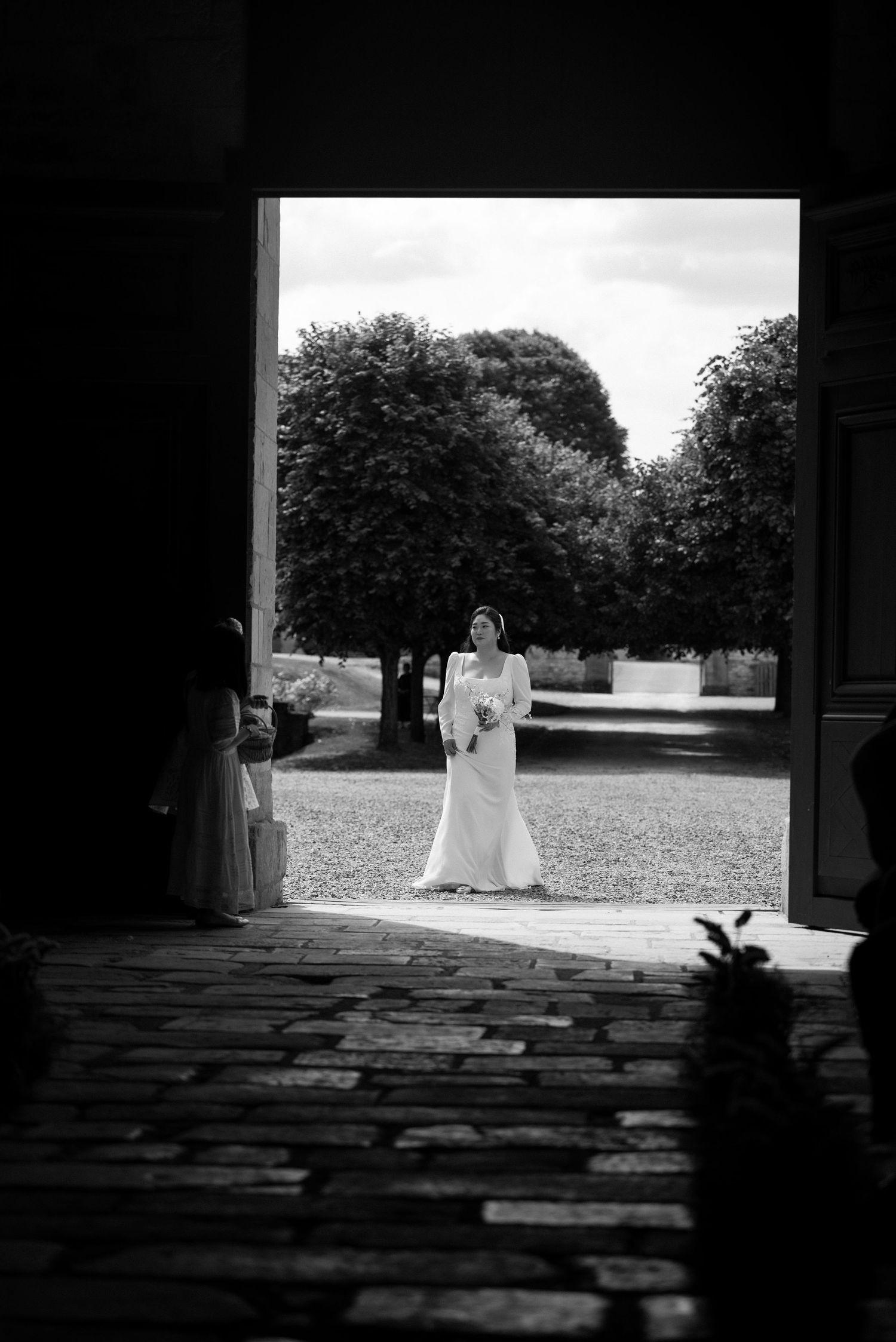 A bride in a white dress stands in a doorway overlooking a garden with trees in the background.