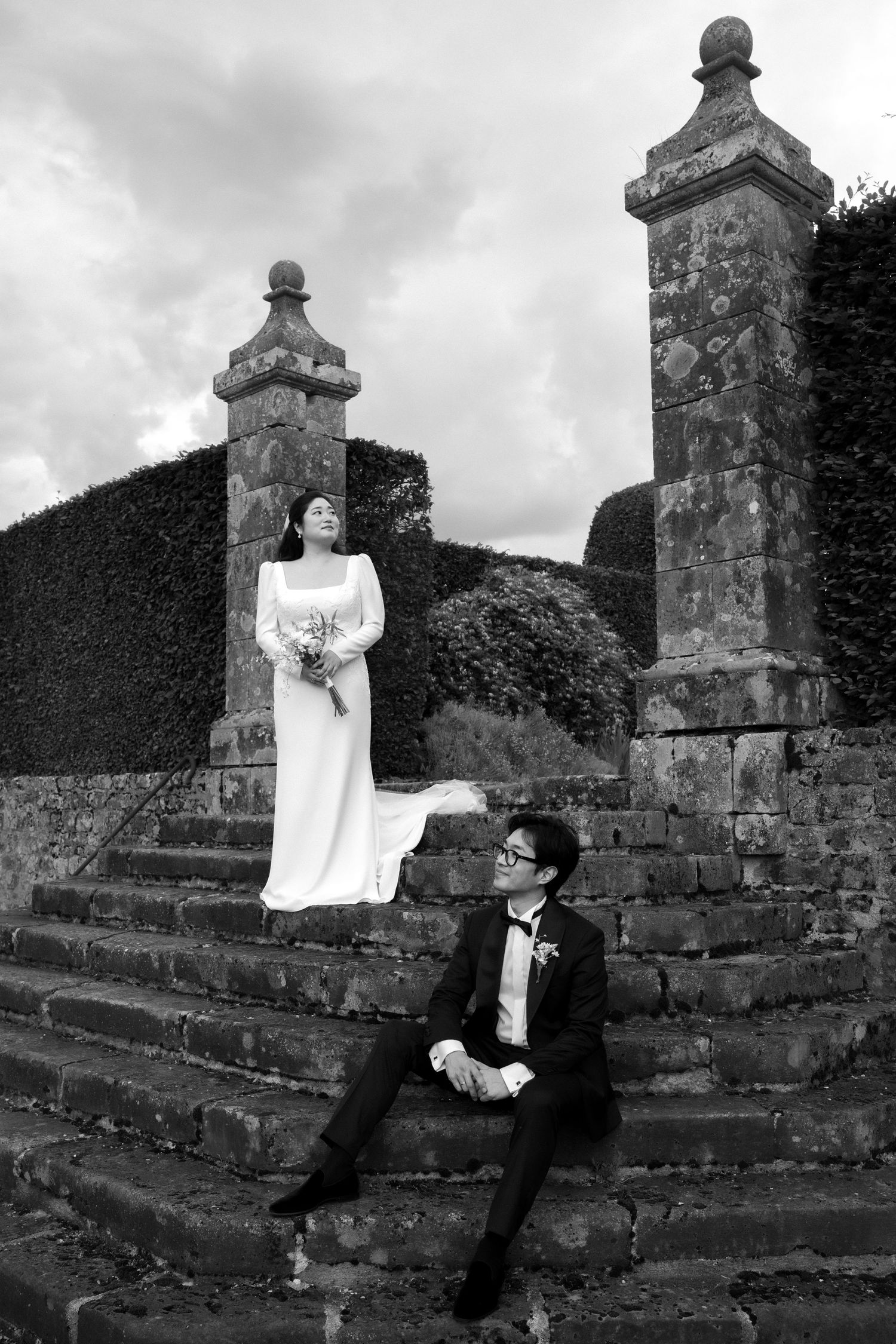 A couple in wedding attire sitting on stone steps against a dramatic cloudy backdrop.